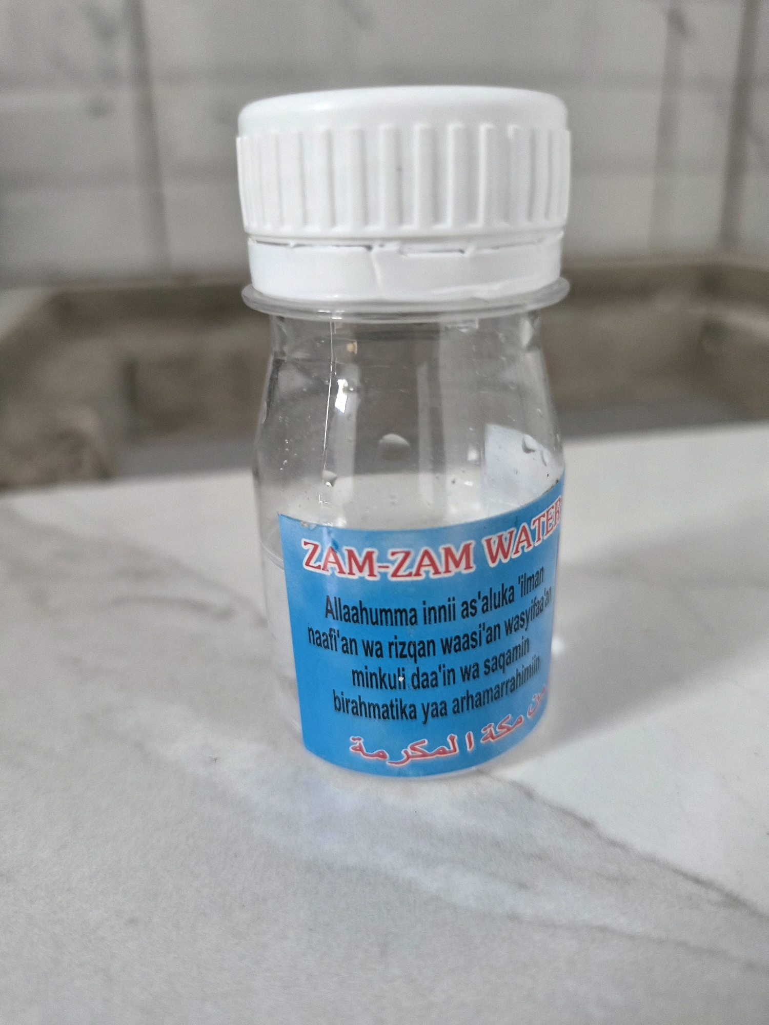 The image shows a close-up view of a small, clear plastic bottle filled with a light-colored liquid, likely Zamzam water. The bottle has a white ribbed cap. A blue label is affixed to the bottle, featuring text in both English and Arabic. The English text reads "ZAM-ZAM WATER" in red and white, followed by a prayer in black. The Arabic text, in red, is partially visible at the bottom of the label. The bottle is placed on a light-colored, speckled surface, possibly a countertop, with a blurred background of what appears to be a tiled wall or backsplash. The lighting is even, and the focus is sharp on the bottle and its label. There are no people visible in the image. The image is a product shot, intended to showcase the bottle of Zamzam water. The location in Srimahi, Indonesia is not depicted or implied by the visual elements of the photograph.