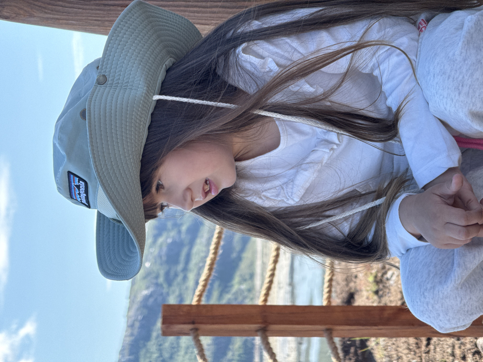 A young girl wearing a light blue Patagonia hat and a white ruffled shirt sits in front of a blurred background of mountains, water, and a wooden railing with rope. The girl's long brown hair falls over her shoulders, and she is looking to her right. The sun is shining, casting a warm light on the scene. The location is the coastal area of Teriberka, Russia, known for its rugged beauty and proximity to the Arctic Ocean. The overall atmosphere is one of peaceful observation and connection with nature.
