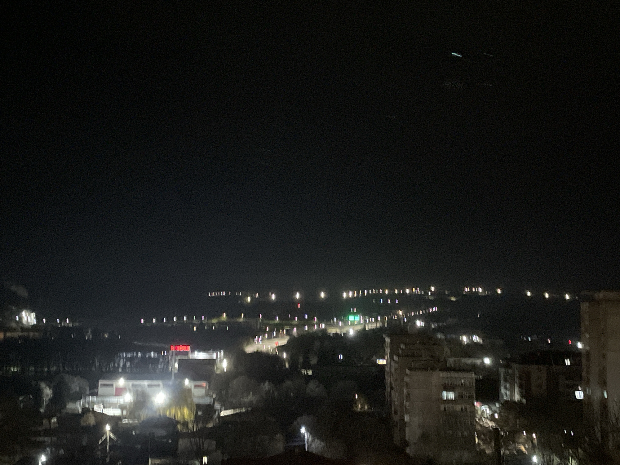 This is a nighttime view of a city, likely Razgrad, Bulgaria, based on visible elements. The scene is dominated by darkness, but city lights punctuate the landscape. In the distance, a string of lights suggests a road or infrastructure stretching along the horizon. A cluster of brighter lights in the mid-ground likely indicates a more populated area, with commercial buildings possibly identified by illuminated signs in red, and residential buildings. The lower part of the image features darker areas, potentially residential neighborhoods, with scattered street lights and building illuminations. The sky is dark. There are no identifiable people. Buildings are the main subjects. No interactions or actions are evident.