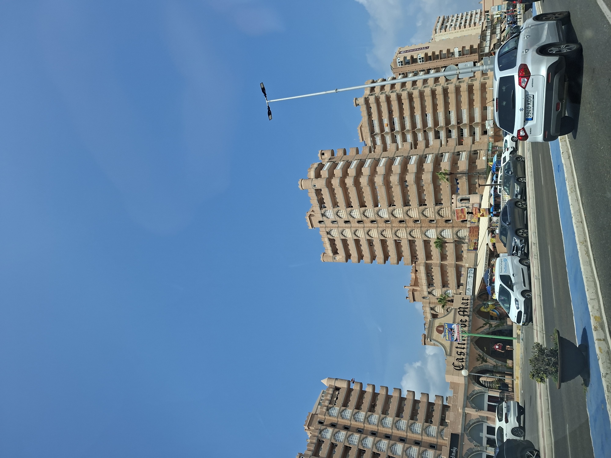 The image depicts a street scene in front of a building complex named "Castillo de Mar" in San Javier, Spain. The buildings are tall, multi-story structures with a distinctive architectural style featuring arched windows and crenellated parapets. They are primarily a light brown or beige color. In the foreground, a road with blue and white markings is visible, with several cars driving on it, including a white Suzuki Vitara with license plate "3940 KDG". There is a potted plant on the left side of the road. The street level features various businesses and shops with awnings and signs, including one that says "MaleniJet" and another that appears to be "Marrakech Bazaar". The sky above is clear and blue with some scattered white clouds. The overall scene suggests a commercial and residential area by the coast.