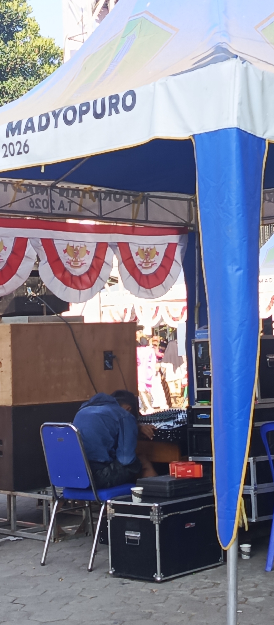 A medium shot captures an outdoor event setup. A blue and white tent with the text "MADYOPURO 2026" is prominent. Beneath the tent, a person wearing a blue jacket is seated on a blue chair, appearing to be working with audio equipment. Various black equipment cases are visible, along with large wooden speaker boxes. Red and white decorative banners, featuring a crest with birds, are hung across the front of the setup. The ground is paved with grey cobblestones, and a leafy green tree is visible in the upper left corner. The lighting suggests a bright, sunny day.