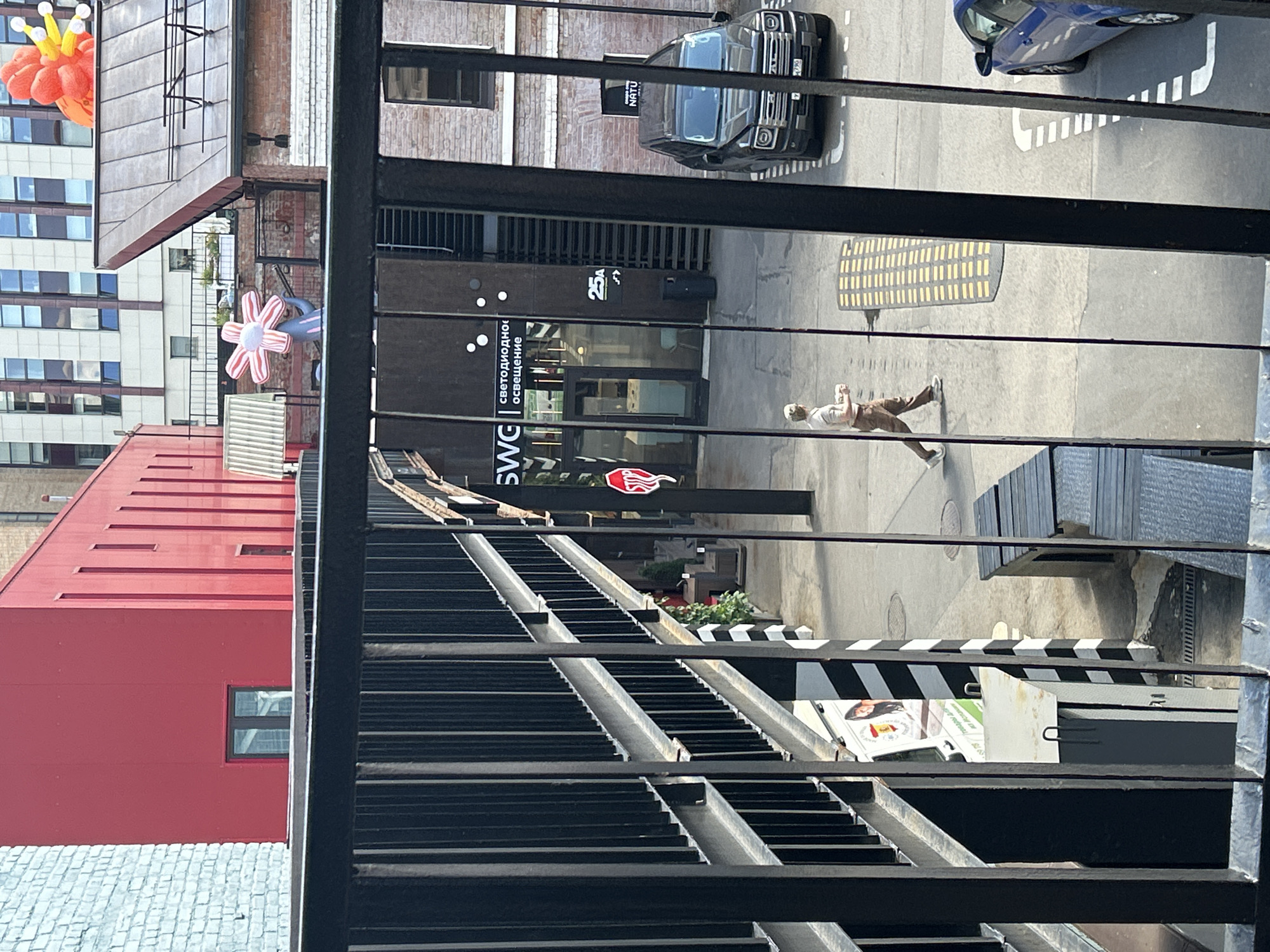 The image shows an outdoor scene with several buildings and a person walking. In the foreground, there is a black metal railing. Behind the railing, a red building is visible on the left, with a light blue brick building next to it. Further back, a dark building with large windows displays the text "SWG" and "светодиодное освещение". To the right of this building, a black SUV is parked. A person wearing a light-colored shirt and brown pants is walking across the paved area, holding a white cup. A blue car is parked to the right of the SUV. In the background, a taller building with many windows is visible, and above a rooftop, an orange inflatable sculpture with yellow and white elements is seen. The ground is paved, and there are yellow speed bumps. The overall environment suggests an urban setting, possibly a courtyard or a street.