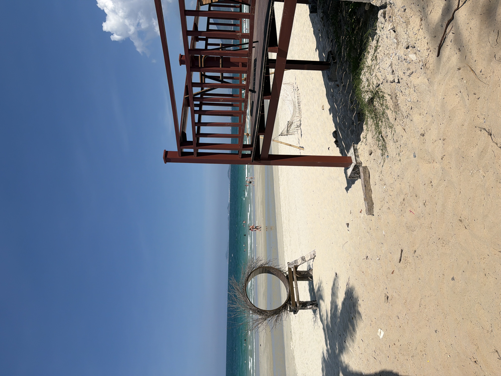 A wide-angle, eye-level shot captures a serene beach scene under a clear blue sky. In the foreground, a sandy beach stretches towards the turquoise ocean, with gentle waves lapping at the shore. To the right, a reddish-brown wooden deck with railings overlooks the beach, and a white hammock is suspended beneath it. On the left side of the image, a circular art installation made of branches rests on a wooden stand. Several people are visible in the water, swimming and wading, while others walk along the shoreline. The overall setting suggests a leisurely day at a coastal resort in Dong Ninh Hoa Ward, Vietnam.