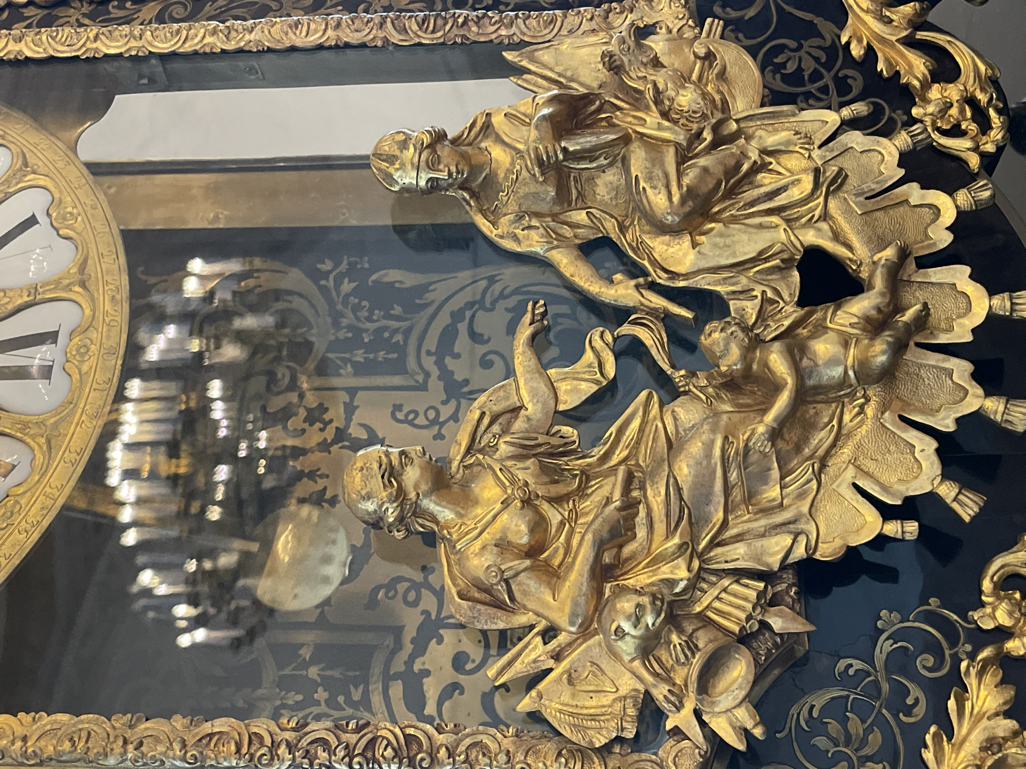 This image is a close-up of a gilded, ornate clock, likely an antique, displaying the numbers 21-35 on its face. The clock is encased in glass and features intricate golden figures in relief adorning its surface. These figures appear to be classical in style, possibly depicting mythological or allegorical scenes. The background is black with a subtle patterned design. Reflections are visible within the glass encasement, suggesting the surrounding environment. Based on the style of the clock and its location, it is plausible that this clock may be found in a museum or historical building in Saint Petersburg, Russia, known for its opulent palaces and collections of decorative arts.