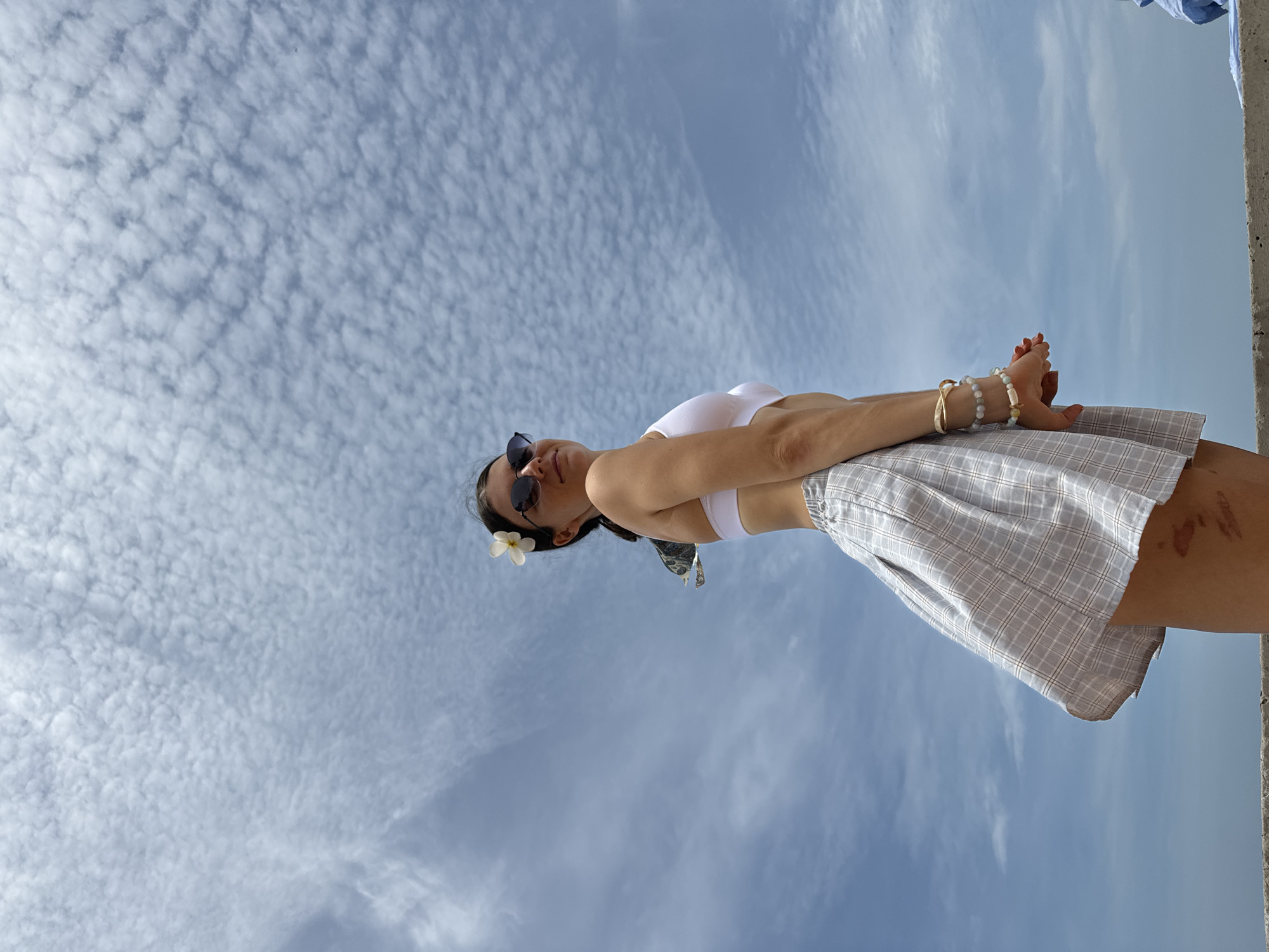 A young woman with dark hair, adorned with a white flower behind her ear, is captured in a low-angle, medium shot. She wears a white bikini top and a light gray plaid skirt, with multiple bracelets on her wrists. She is looking to her right, away from the camera, with sunglasses on. The background is a vast sky filled with dappled clouds. She is positioned on a concrete surface. The image lacks any discernible landmarks or contextual clues to pinpoint a specific location within Phu Thuy Ward, Vietnam.