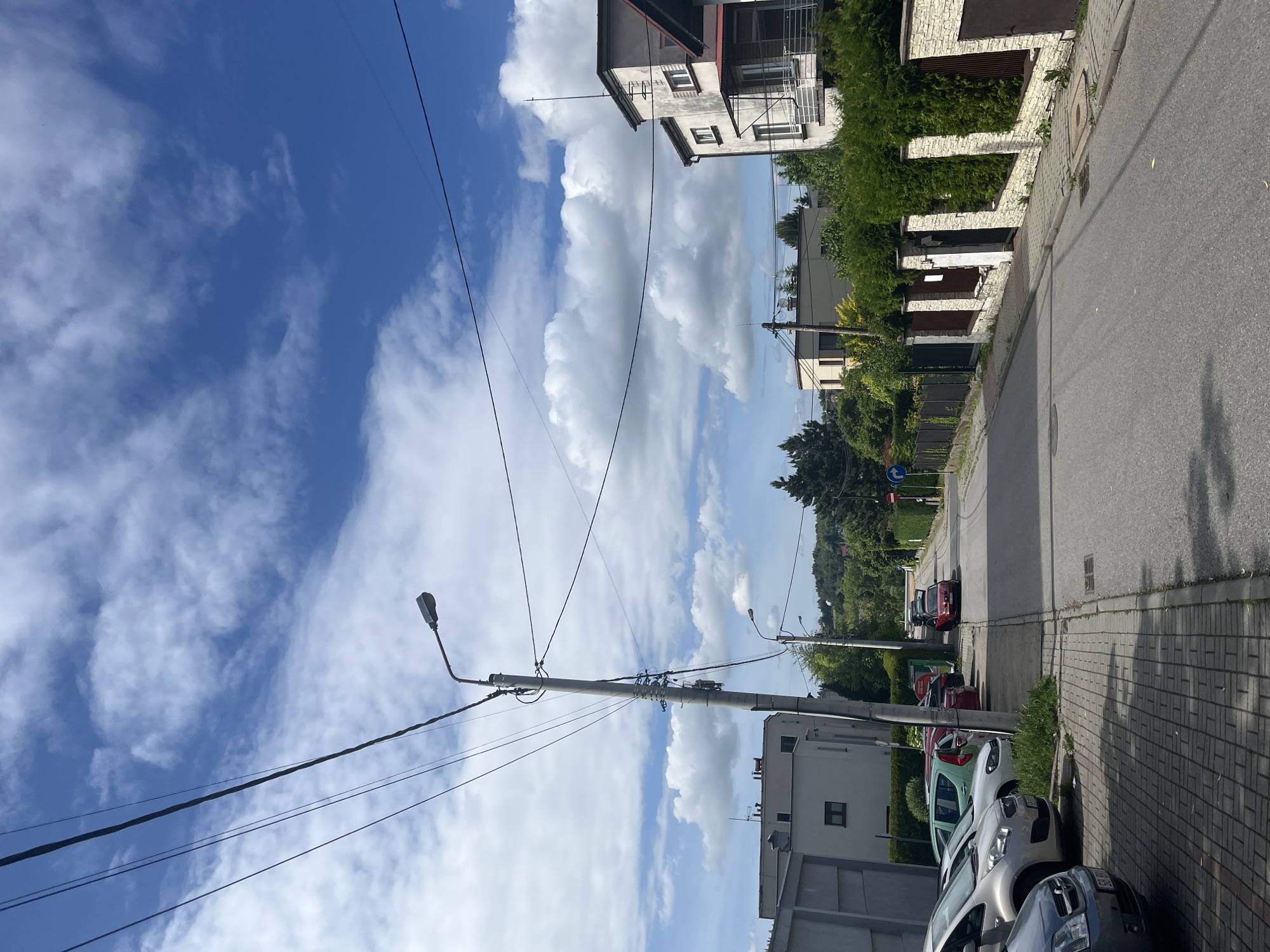 This daytime photograph captures a residential street under a bright, partly cloudy sky. The image, when viewed in its correct vertical orientation, shows a street extending into the distance, appearing to curve to the right. On the left side of the street, a row of cars is parked along a paved sidewalk, with vehicles ranging in color from silver to light green and red. A section of a multi-story building is visible behind the parked cars and sidewalk.
On the right side of the street, a series of residential properties are lined with fences featuring light-colored stone or brick columns and dark panels, topped by lush green hedges and foliage. Parts of several houses can be seen above these green barriers. Overhead, multiple utility poles with numerous electrical wires and streetlights crisscross the scene, indicating an urban or suburban setting. A prominent utility pole with a streetlight stands in the mid-ground on the left.
The sky is a vibrant blue, filled with scattered white cumulus clouds. Shadows cast on the asphalt road and paved sidewalk confirm that it is a sunny day. In the background, further down the street, a blue circular road sign with a white arrow pointing right is visible. No people are present in the photograph, and no legible text can be identified on any objects or signs. The scene depicts a calm, ordinary moment in a residential area.
