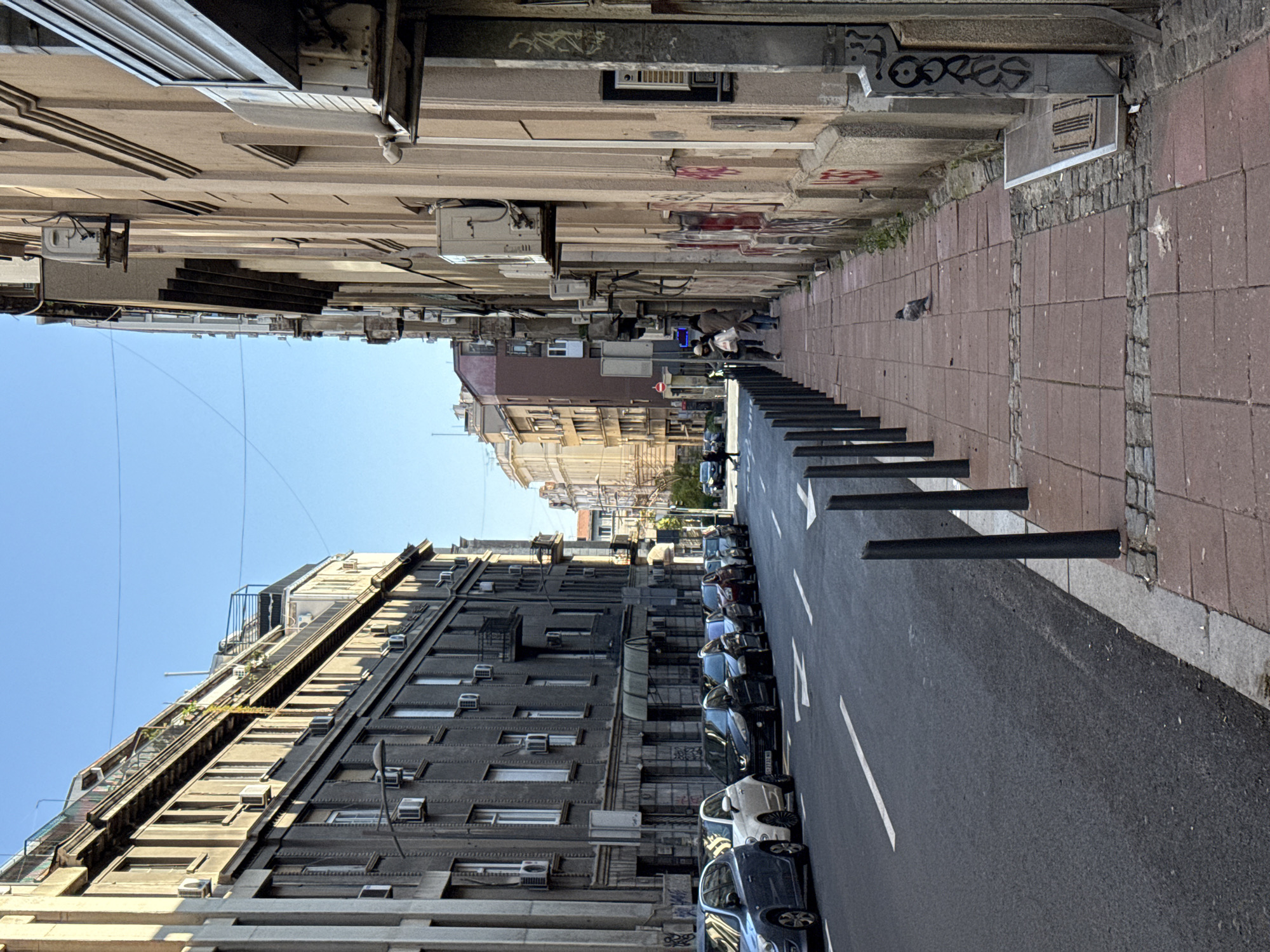 The image captures a street in Belgrade, Serbia. The street is lined with buildings on both sides, varying in height and architectural style. The buildings appear to be older, with signs of wear and tear, including graffiti on the facades. Air conditioning units are visible in numerous windows. Cars are parked along the right side of the street. A pedestrian sidewalk is on the left side of the street, with people walking in the distance. Black bollards line the edge of the street, separating it from the sidewalk. The street is paved, with white lane markings. The sky is blue and clear. A pigeon is visible on the sidewalk near the bollards.