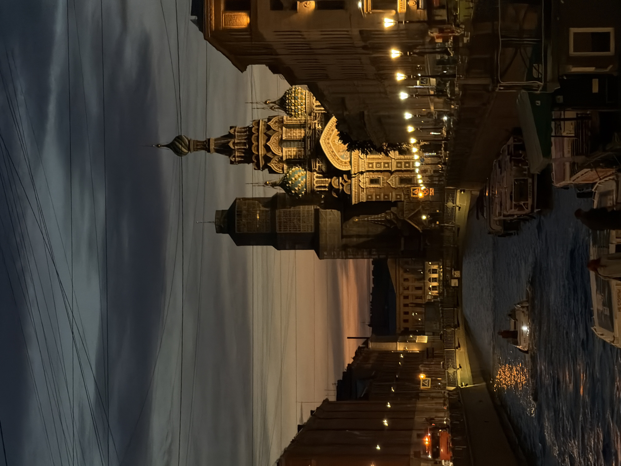 This is a nighttime photograph of the Church of the Savior on Spilled Blood in Saint Petersburg, Russia, taken from a canal. The sky is dark with swirling clouds, transitioning to a warm orange glow near the horizon. The church's distinctive onion domes and ornate facade are illuminated, catching the light. Buildings line both sides of the canal, with warm yellow lights emanating from windows and streetlamps. Several boats are visible on the dark, rippling water of the canal. One boat in the foreground appears to be moving, creating a wake. Other boats are docked along the canal's edge, some covered with tarps or awnings. A few people are visible on the boats and on the walkways. Overhead, a complex network of power lines crisscrosses the sky, adding a layer of urban texture to the scene. The overall impression is one of a historic European city at dusk, with its iconic architecture set against a dramatic sky and illuminated by artificial light.