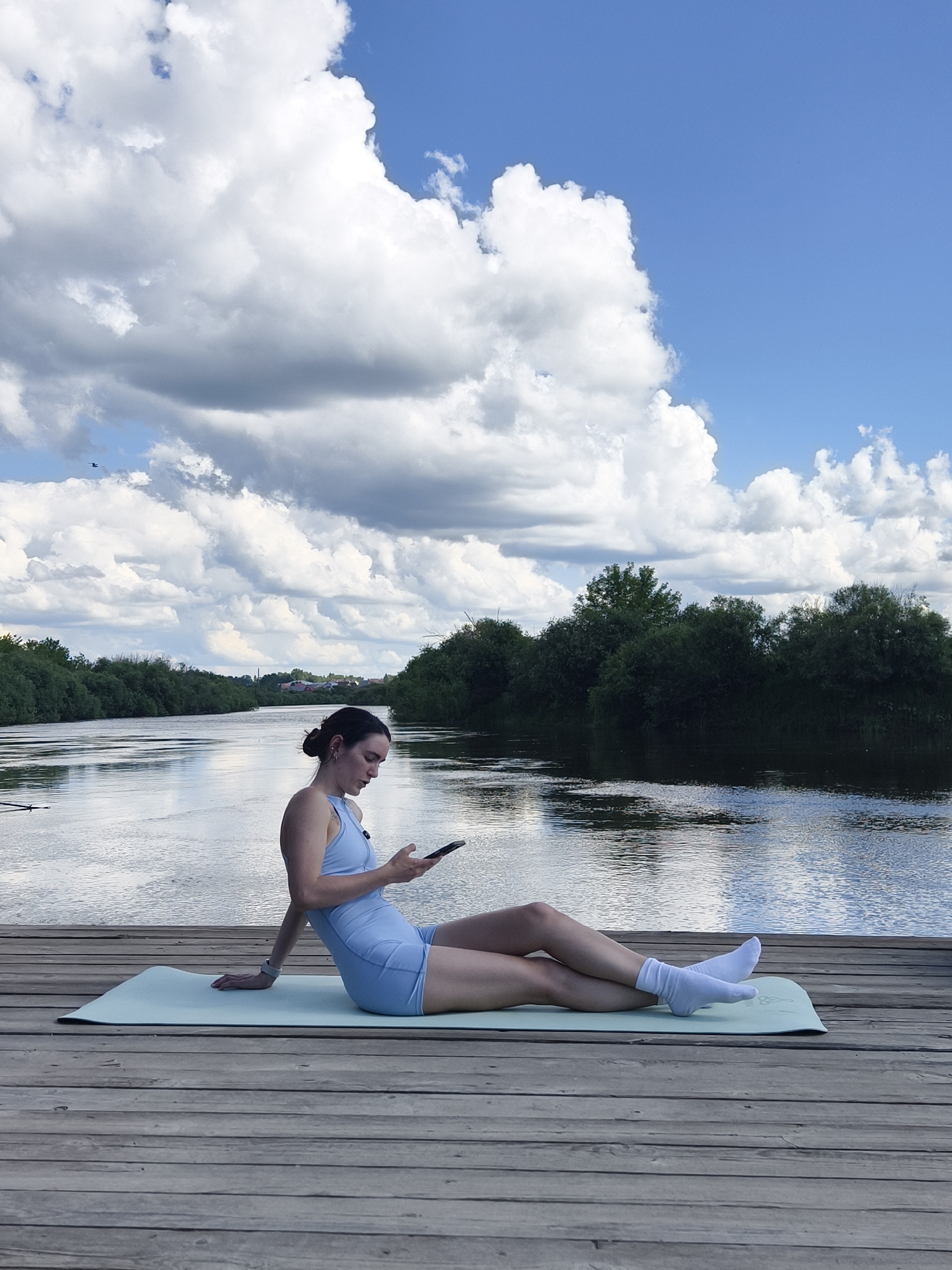 A woman is sitting on a yoga mat on a wooden pier, looking at her phone. She is wearing a light blue athletic outfit and white socks. The pier extends over a calm body of water, likely a river, with lush green trees lining the opposite bank. The sky above is a vibrant blue with large, white cumulus clouds. In the distance, along the riverbank, there are hints of buildings and more trees. The scene appears to be a peaceful, outdoor setting, possibly in or near the city of Onochino, Russia, given the context provided.