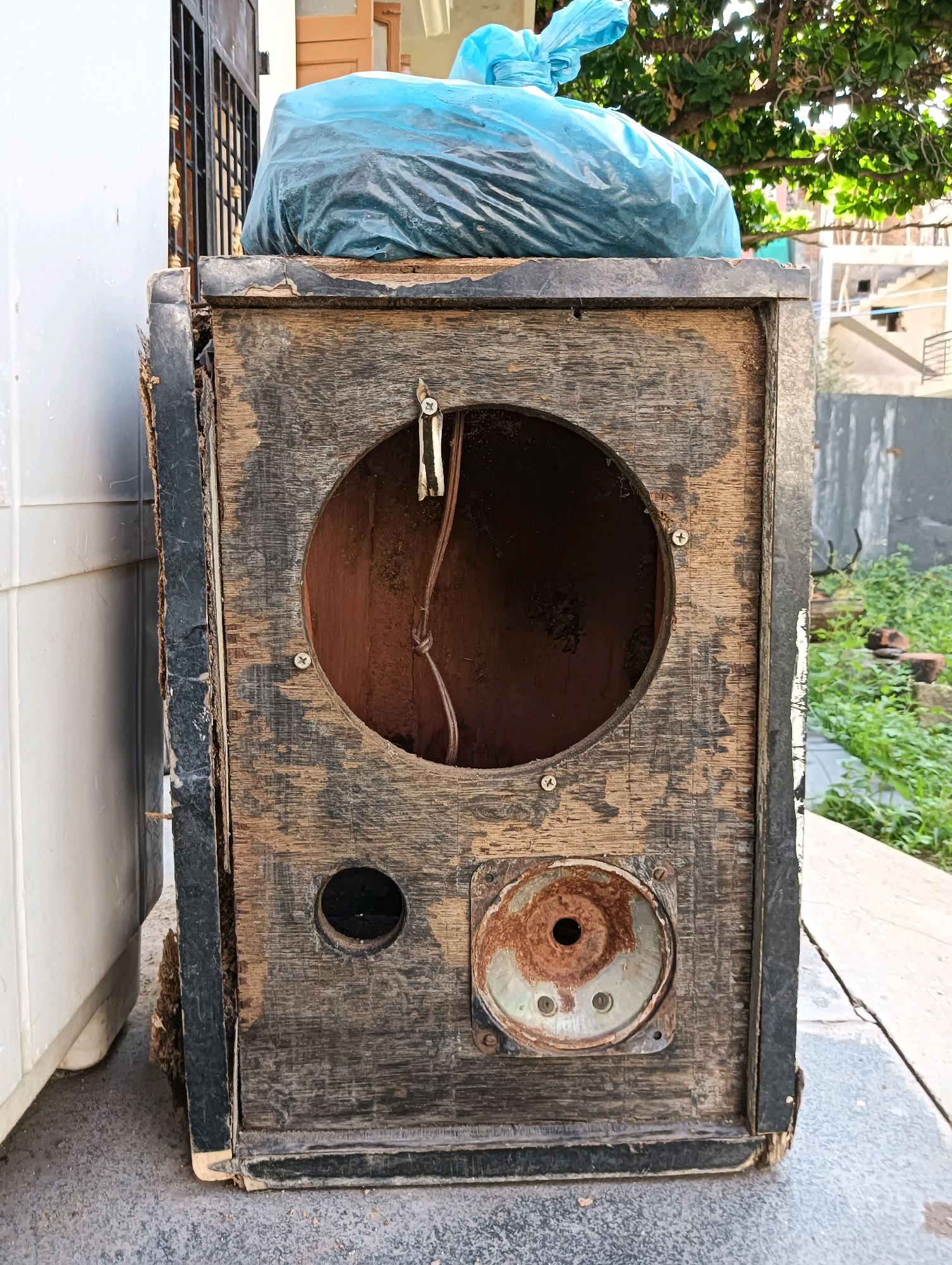 An old, weathered wooden speaker cabinet rests on a paved surface in front of a white appliance and a metal gate. The speaker cabinet is damaged, with chipped paint and exposed wood. It has a large circular opening for a speaker, a smaller circular hole below it, and a rusted circular metal component. A blue plastic bag filled with trash sits on top of the cabinet. The background shows green foliage and part of a building. The scene is located in Hosapete, India.