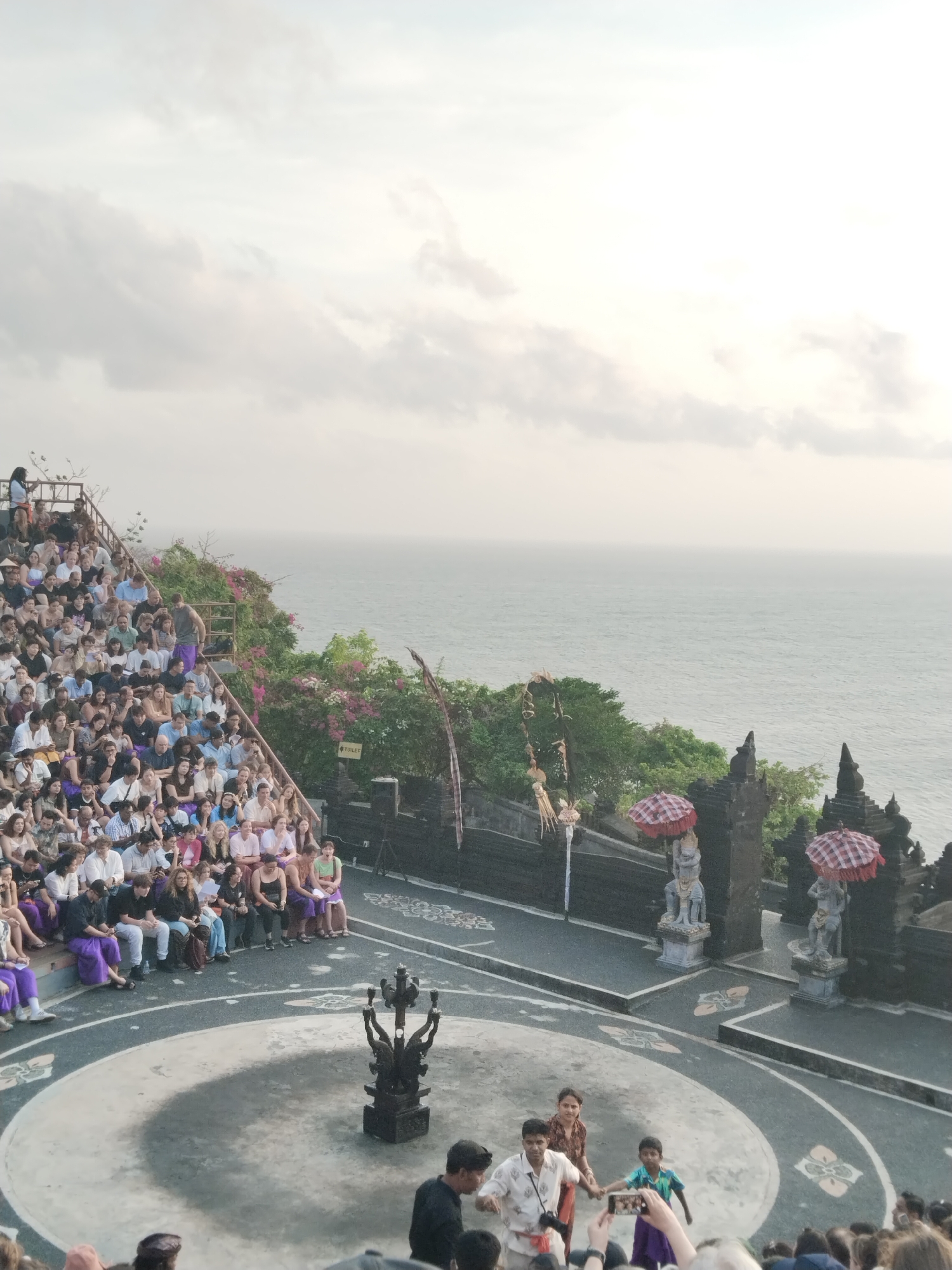 In Kuta Selatan, Indonesia, an outdoor amphitheater overlooking the ocean hosts a cultural performance. A large audience is seated on tiered stone bleachers, many facing a circular performance area. In the center of the performance area, a dark, ornate statue stands on a pedestal. At the rear of the performance area, a gateway with two stone figures holding patterned umbrellas marks an entrance. Performers, including a man and a child in traditional attire, are present in the performance space, interacting with each other. The sky is overcast, and the sea is visible in the background.
