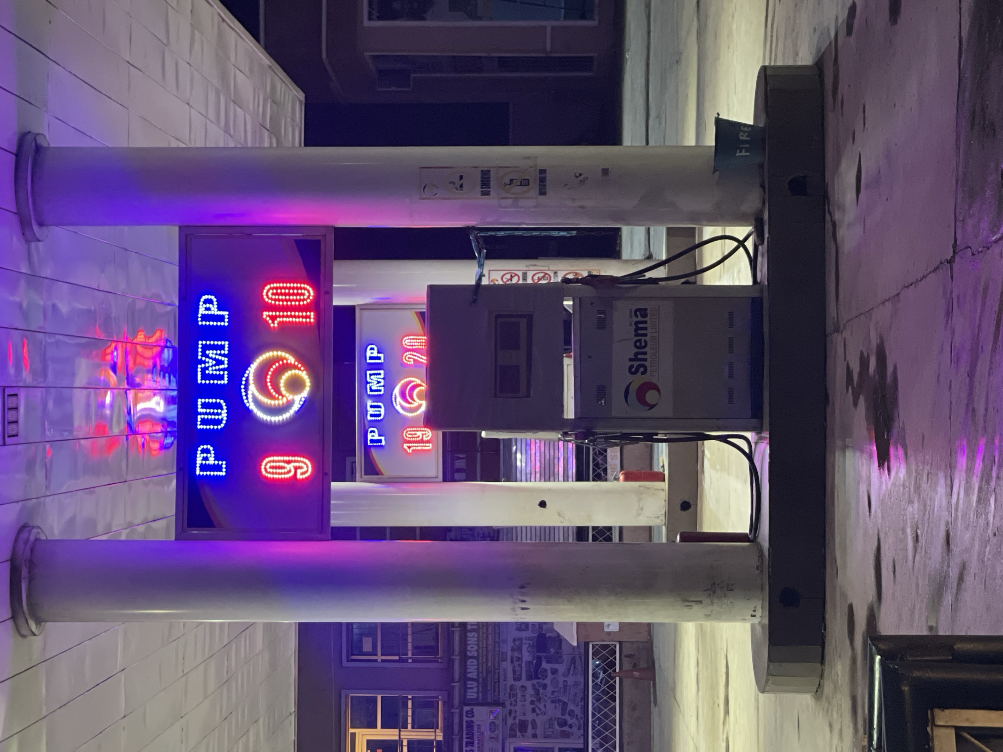 The image shows a gas station, presumably in Hayin Banki, Nigeria. Two illuminated signs indicate prices: "PUMP 910" and "PUMP 1920." A gas pump unit, labeled "Shema Petroleum Limited," is positioned between two white support columns. A "FIRE" sign is visible at the top of the frame. Below, a storefront featuring the name "ULU AND SONS TI" and "TRADING CO." is visible. Additional windows and what appears to be a promotional display are visible, adding context to the urban setting. The photograph is taken from a lower angle, giving prominence to the gas pump and signs.