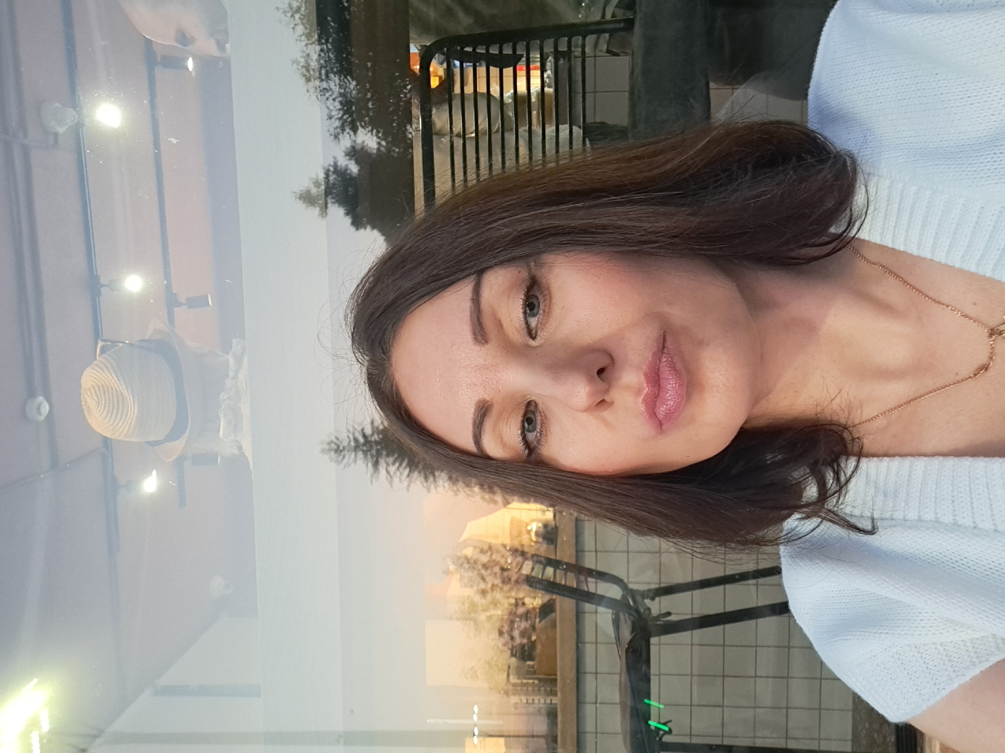A medium shot shows a woman with brown hair and fair skin, looking directly at the camera with her lips pursed. She is wearing a white knitted sweater and a delicate gold necklace. The background is blurry and appears to be a reflection in a window, showing a hat, lights, and a partial view of a room with chairs. The scene is likely indoors or near a window with a view of the outdoors, and it is not possible to determine the specific location within Irkutsk, Russia from the image alone.