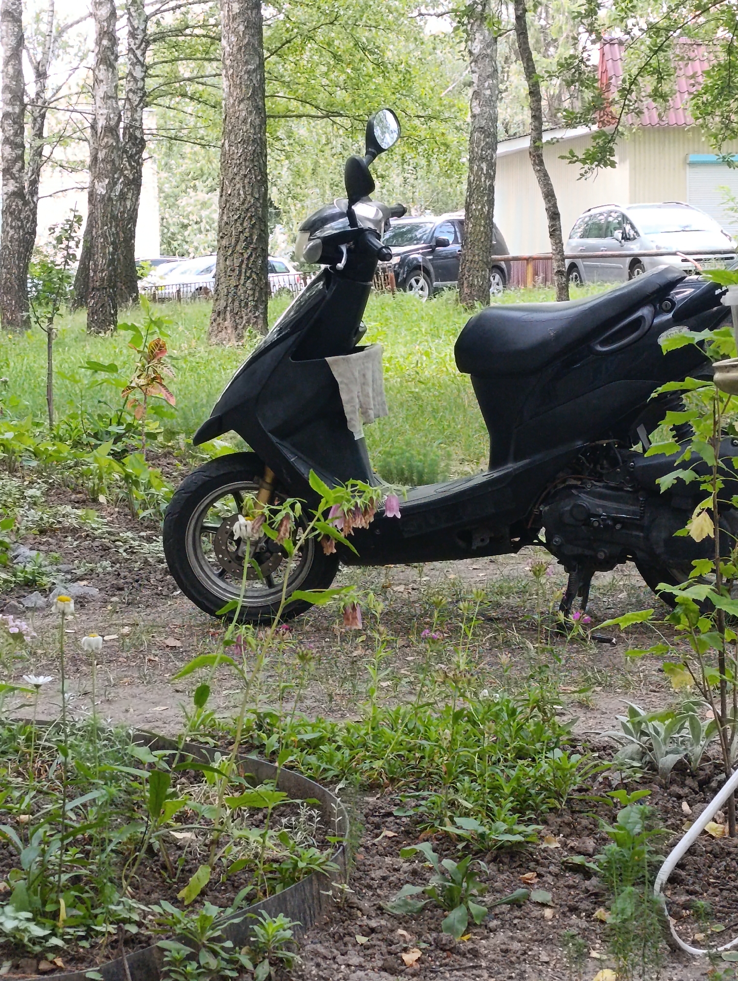 A black scooter is parked on a dirt path in a grassy area with trees. In the background, a light-colored building with a red-tiled roof is visible, along with several parked cars. The scene appears to be in a residential or park area. This image was captured in the city of Ladyzhyn, Ukraine.