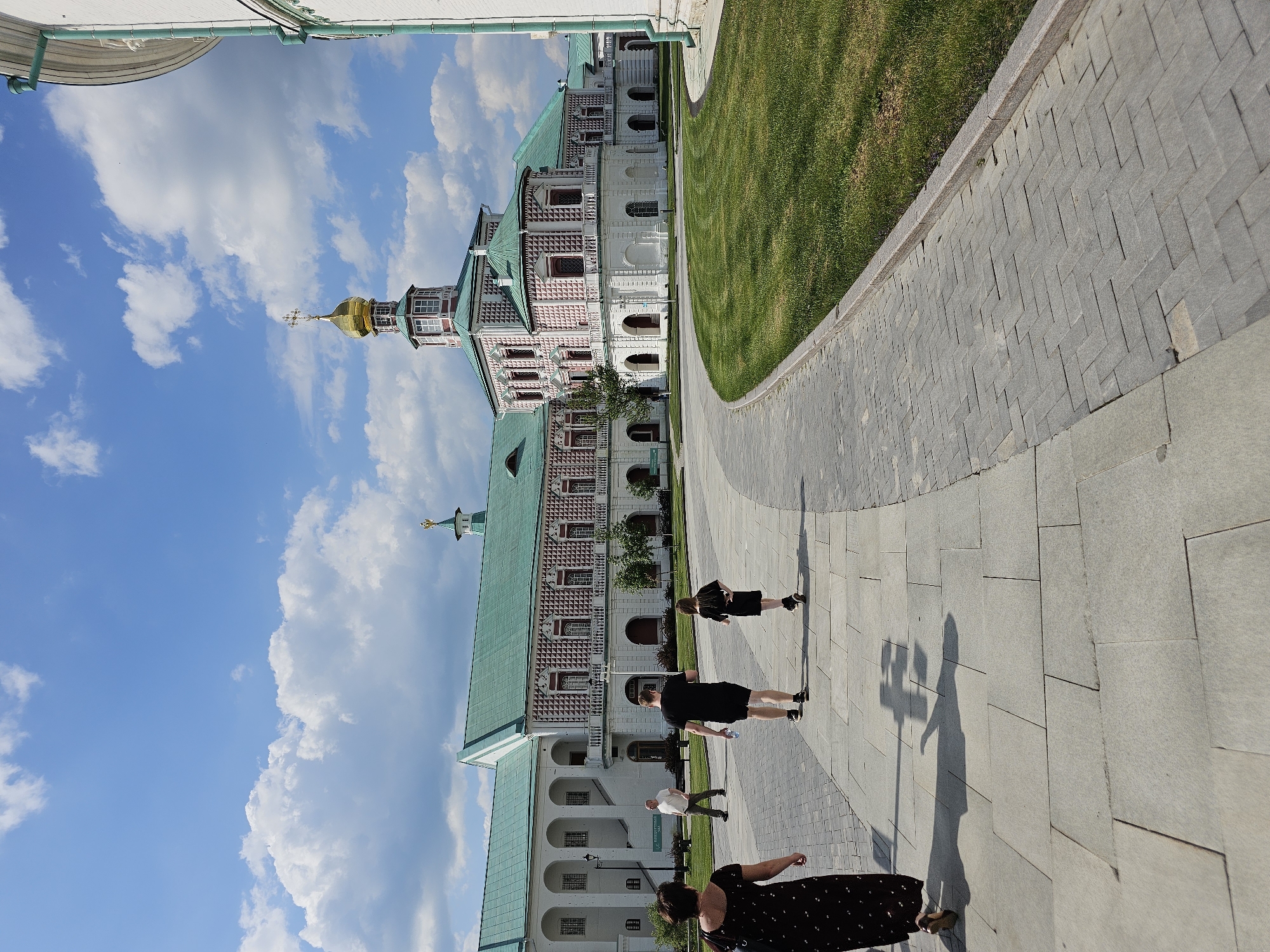 The image shows the New Jerusalem Monastery in Istra, Russia. The monastery complex is a mix of white buildings with red brick detailing and green roofs. A golden dome with a cross is visible. People are walking on a paved pathway that curves next to a grassy area. The sky is blue with fluffy white clouds. Shadows of the people are visible on the pavement.