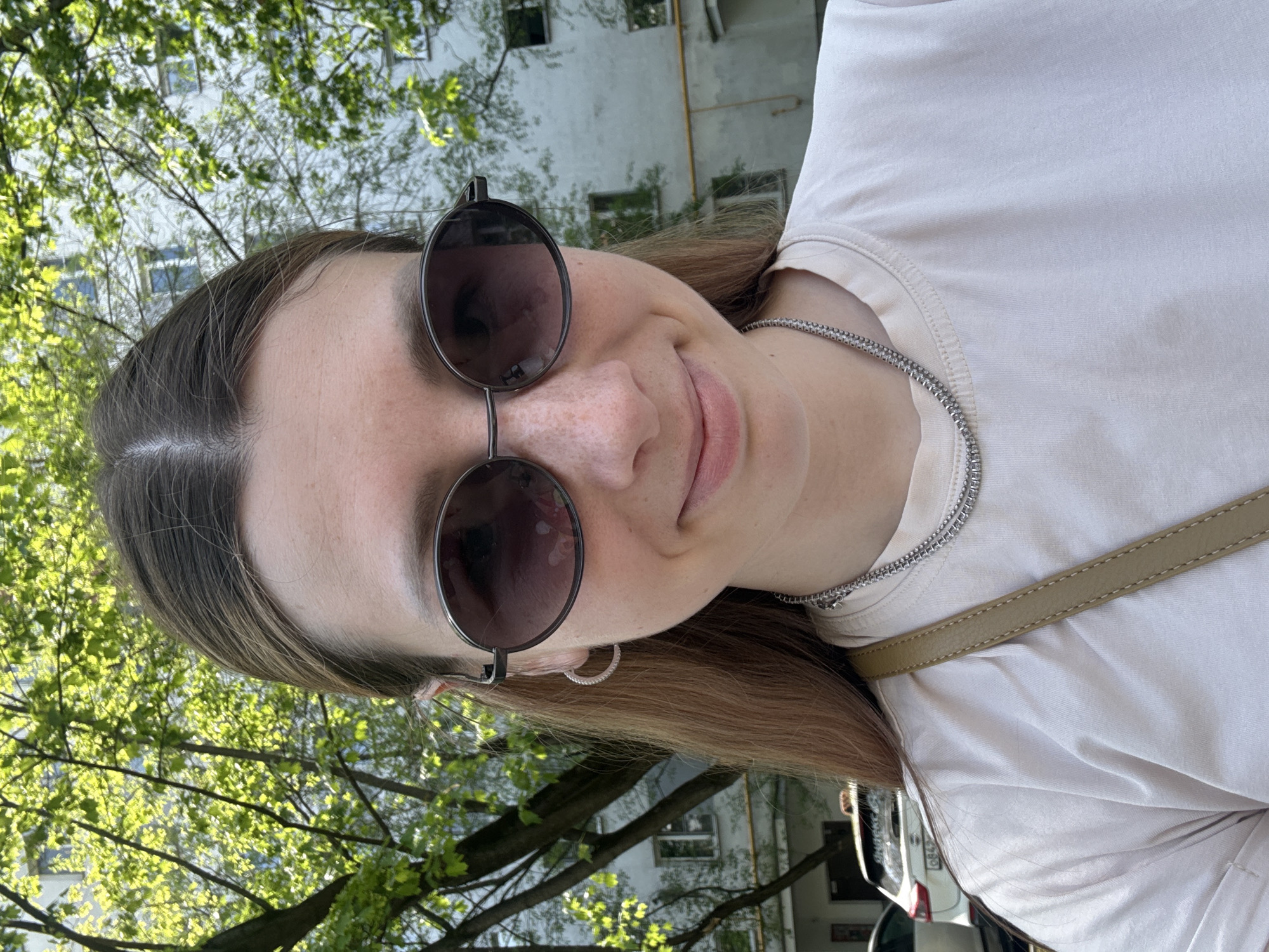 A woman with long brown hair and freckles is wearing round sunglasses and a silver chain necklace, posing outside on a sunny day. Behind her are green trees and a building with windows, suggesting an urban park setting. Based on architectural style, it is likely that the location is in Moscow, Russia.