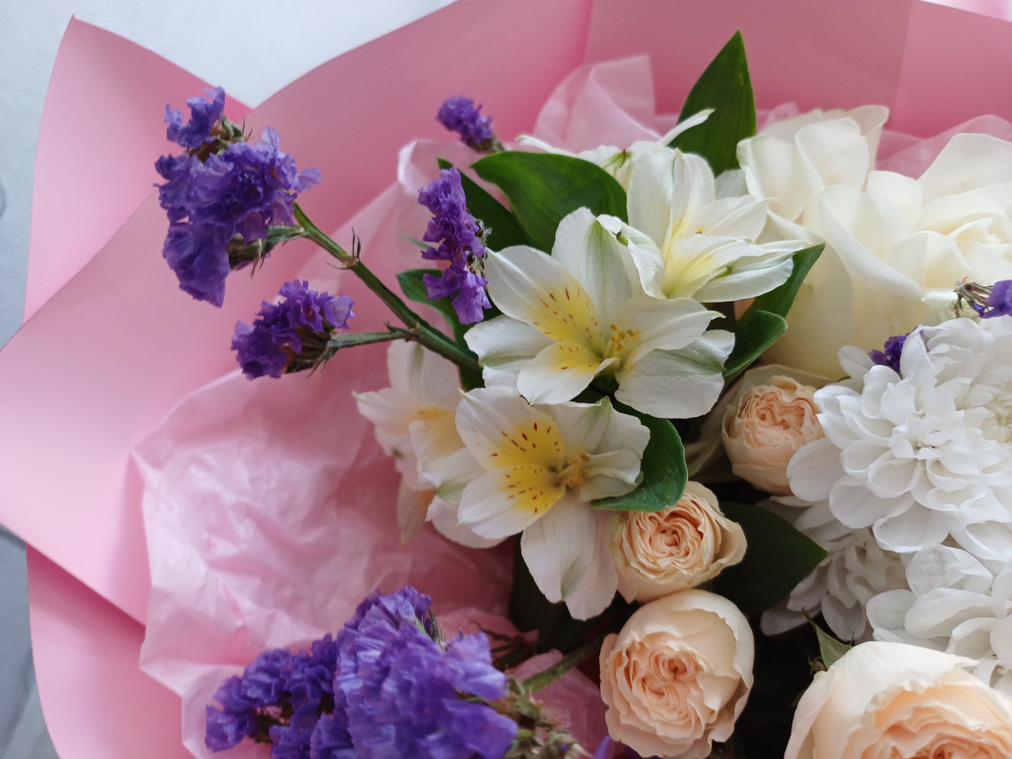 A close-up shot captures a bouquet of flowers, likely wrapped in pink paper. The bouquet features a variety of blooms in different colors and textures. Prominent white alstroemeria with yellow centers are visible, interspersed with delicate, light pink roses. Clusters of small, purple flowers, possibly statice, add a vibrant contrast to the softer hues. White chrysanthemums, with their layered petals, also contribute to the bouquet's fullness. The background is a soft pink, suggesting the bouquet is either presented against a pink surface or wrapped in pink paper. The lighting is bright, highlighting the details of the flowers and their colors.