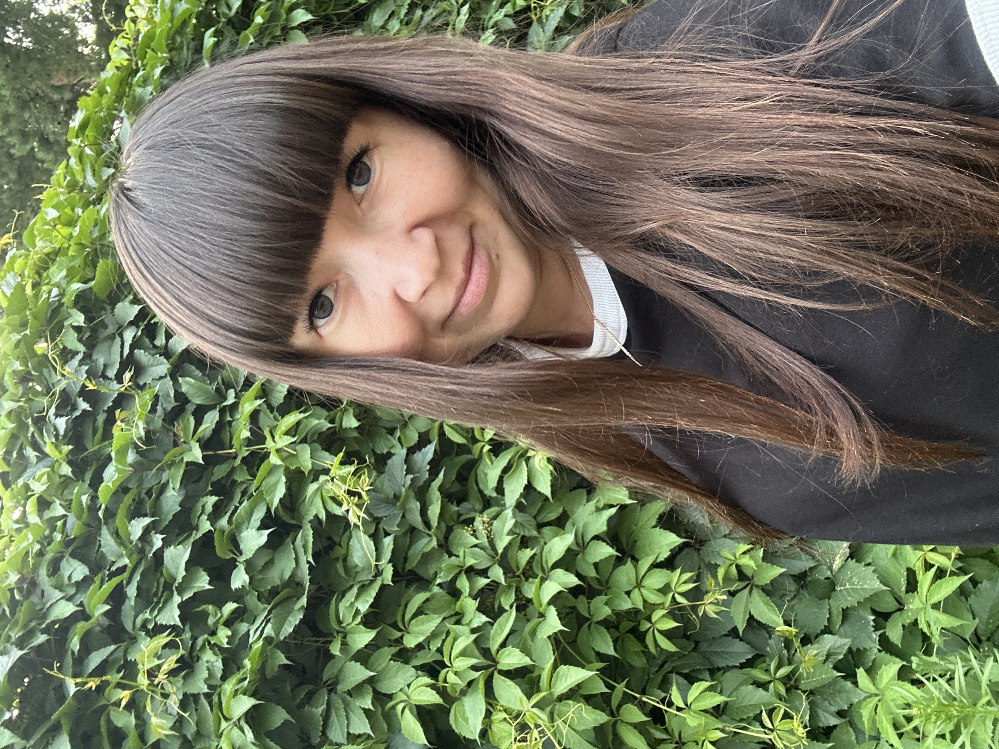 A woman with long brown hair and bangs, wearing a black sweatshirt with a white collar, stands in front of a dense green wall of ivy. The woman is smiling and looking at the camera. The background is a lush green wall of ivy, with some trees visible in the upper right corner. The lighting is natural and appears to be daylight. There is no visible indication of the specific location within the city of Noviy, Russia.