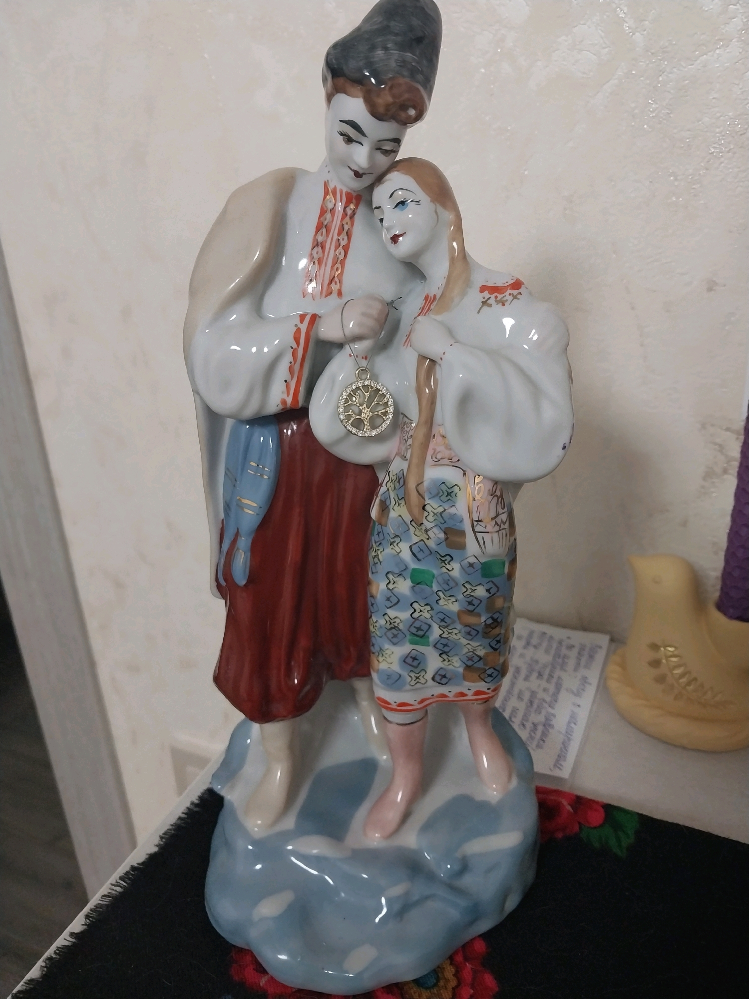 A ceramic figurine of a couple in traditional Ukrainian attire stands on a blue, cloud-like base. The male figure, on the left, wears a dark hat, a white shirt with red and gold embroidery, and a red skirt. He holds a blue pouch at his side. The female figure, on the right, has long blonde hair and wears a white top with red embroidery and a patterned blue skirt. She has a white bag slung over her shoulder. A gold circular pendant with a tree-like design is suspended between them. The figurine is placed on a surface with a black, fringed cloth and a small yellow bird figurine is visible to the right, behind the couple. A piece of paper with handwritten text is also present behind the figurine. The background is a textured, light-colored wall. The scene appears to be indoors, likely in a domestic setting within the city of Dnipro, Ukraine.