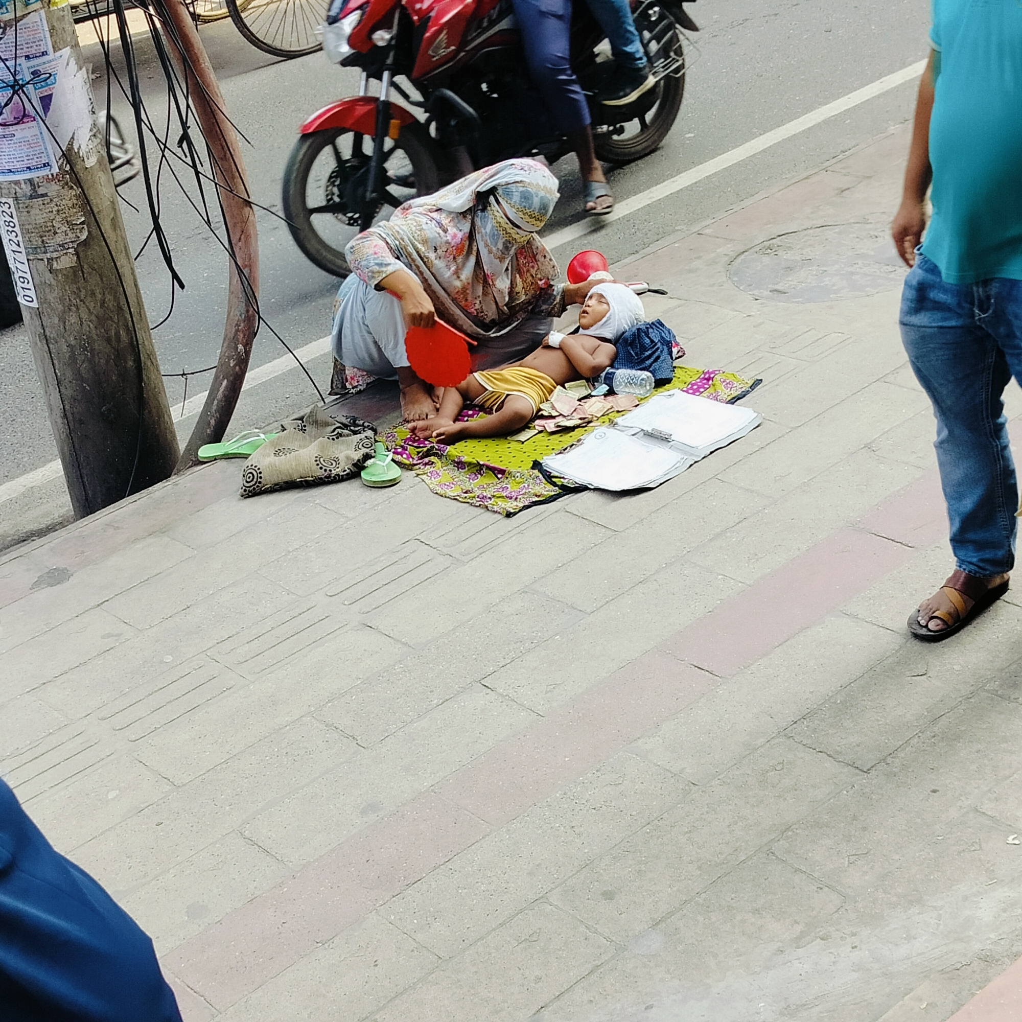The image shows a woman sitting on a patterned cloth on a sidewalk next to a utility pole, near a street with passing vehicles. She is holding a red fan and appears to be fanning a child lying on the cloth. The child has a white cloth wrapped around their head. There are some scattered items on the cloth including money, clothes, and a water bottle. A man in jeans and sandals is standing nearby. The location appears to be urban.