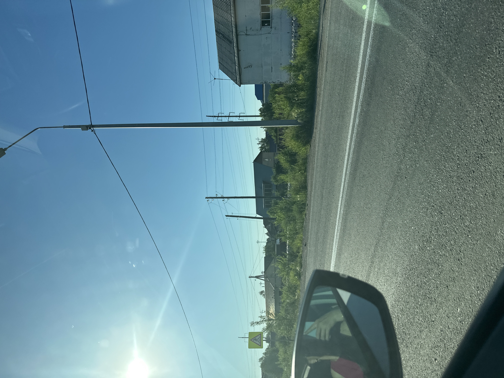The image depicts a roadside scene from a car's perspective, likely in Abakan, Russia. The viewpoint is from inside the vehicle, with the side mirror in the foreground reflecting a person's arm and hand. The scene is bathed in bright sunlight, indicating it is daytime. The sky is a clear, vibrant blue with some lens flares and reflections on the car's windshield.
The road itself is asphalt, with double white lines marking the lanes. To the left of the road, there is a sidewalk or verge covered in green grass and vegetation. Beyond the vegetation, several buildings are visible, some appearing to be industrial or commercial structures with corrugated metal roofs. Power lines and utility poles are a prominent feature, stretching across the frame. In the distance, more buildings and trees can be seen.
A yellow triangular road sign with black symbols is visible on a post, which appears to be a warning sign. The overall impression is of a rural or semi-urban environment on a sunny day. The presence of the road, buildings, and power lines suggests a developed area. The quality of the light and the clear sky point to fair weather.