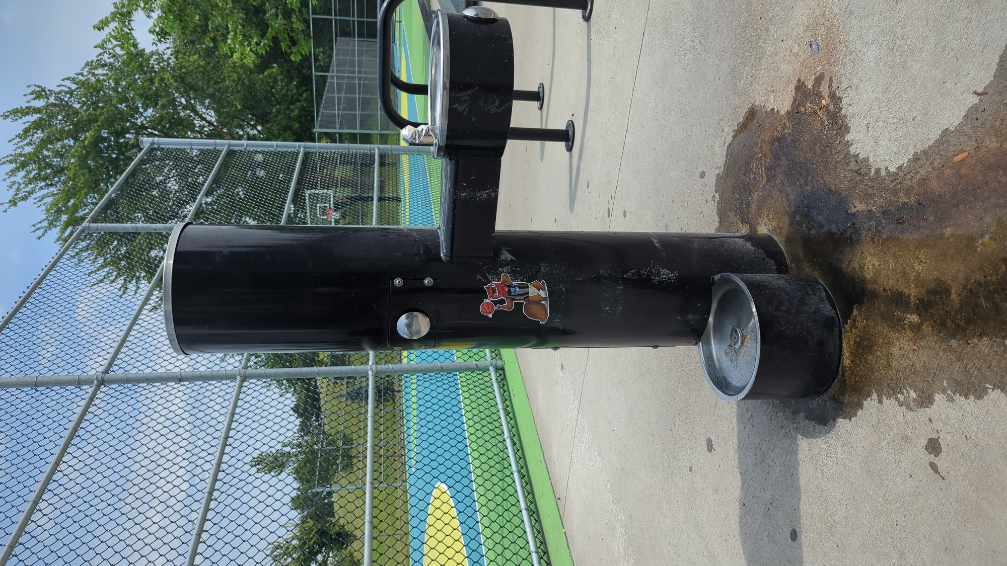 The image shows a black water fountain with a separate dog bowl at its base. The water fountain has a sticker of a cartoon squirrel holding a basketball on its front. Behind the water fountain, there is a chain-link fence and a basketball court with colorful markings in green, blue, and yellow. Trees and a clear blue sky are visible in the background. The ground is concrete, with a wet stain spreading from the base of the water fountain, indicating recent use. The scene appears to be an outdoor recreational area, likely a park or sports complex. No people are visible in the image. The location in Montreal, Canada cannot be determined from the image alone.