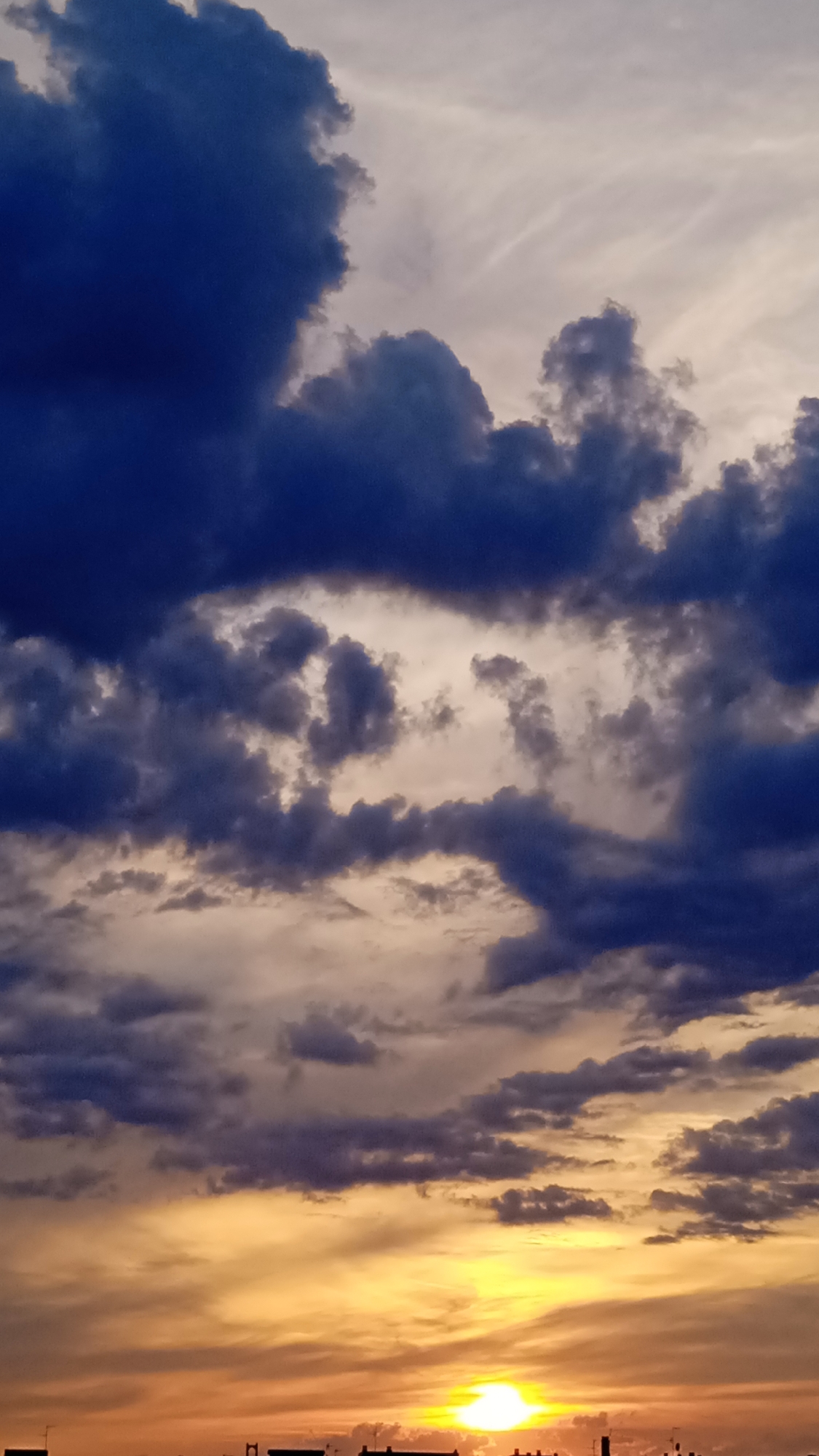 The image depicts a dramatic sky filled with dark, heavy clouds tinged with blue and purple, juxtaposed against lighter, wispy clouds. The lower portion of the sky transitions into a vibrant sunset, with intense orange and yellow hues. Silhouetted against the horizon are the faint outlines of buildings and structures, suggesting an urban setting. The sun itself is a bright, concentrated orb near the bottom right of the frame.