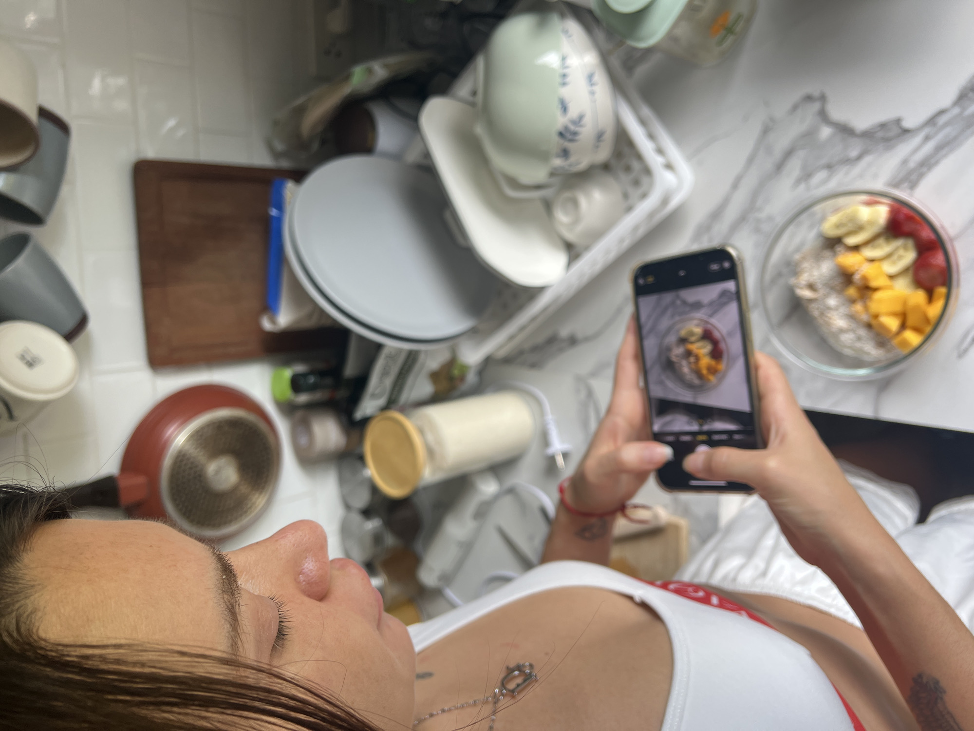 A person is photographing a bowl of fruit and granola on a counter. The counter is marble. A phone is being held in both hands. The person is wearing a white tank top. Behind the bowl are dishes, a cooking pan, and a storage container. The dishes and bowl suggest a kitchen. The exact location is not discernible from the image, only the country of Thailand has been provided.