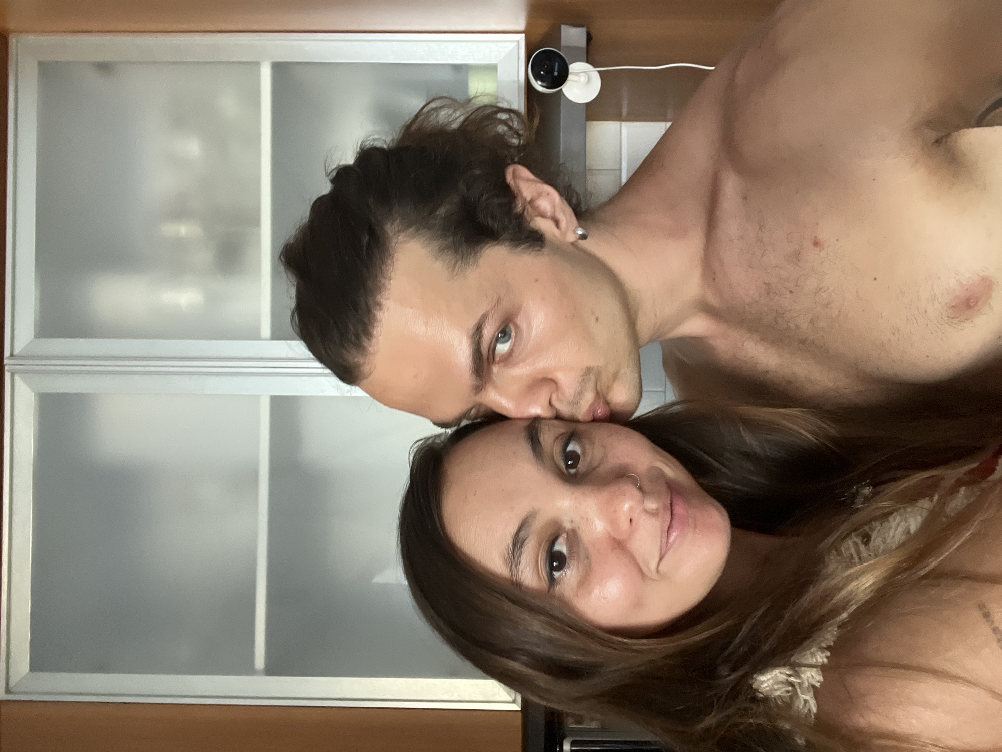 A horizontal medium shot shows two individuals indoors. The man, shirtless, has light skin, wavy dark hair pulled back, and blue eyes. He is kissing the forehead of the woman. The woman has long, light brown hair and a nose ring. Behind them is a wall with a set of white cabinets, and a security camera is mounted above. The location in the city of Mengwi, Indonesia, is not evident in the image.