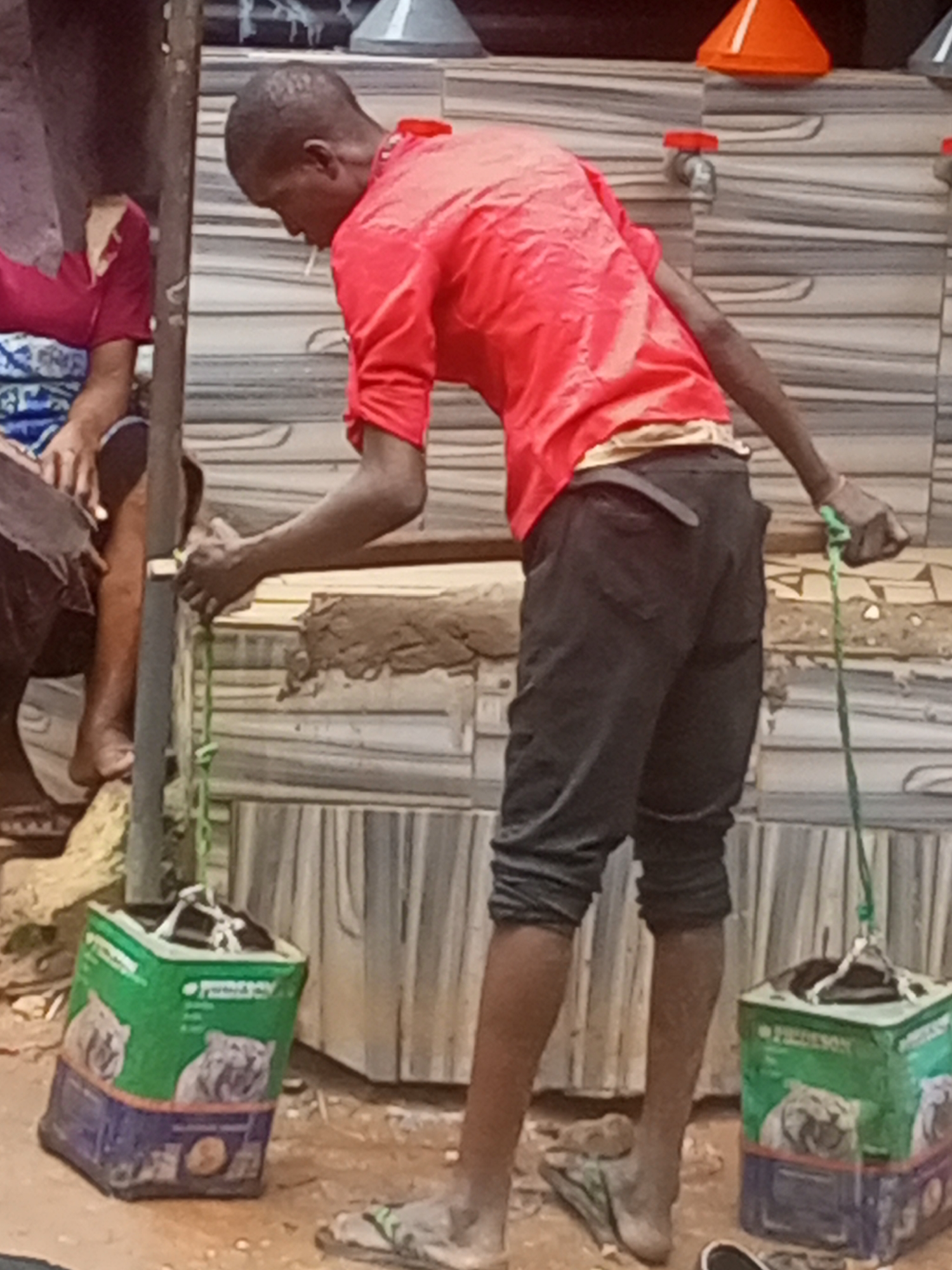 This media file captures a scene of daily life centered around water collection in Onitsha, Nigeria. The main subject is a young male with dark skin and short hair, wearing a bright red short-sleeved shirt and dark pants. He is seen with a cigarette in his mouth and appears focused on the task at hand. To his left, partially obscured, another person with dark skin is seated, wearing a patterned blue and white garment and a red top.
The primary activity depicted is the collection of water. The young man is holding a makeshift handle made of green rope, attached to a repurposed square container. Two such containers are visible on the muddy ground. These containers appear to be recycled plastic jerrycans, featuring green and purple/blue sections. The word "PEDIGREE" is visible on the green part of the containers, alongside images of dogs, suggesting they were originally packaging for dog food.
The setting is an outdoor or semi-outdoor area, characterized by a distinctive striped wall in the background, likely tiled or painted. Several water taps are mounted on this wall; one is notable for its orange conical cap, and another has a red circular cap. There is some built-up mud or earth along the base of the wall. The ground itself is dirt and mud, and flip-flops are visible. The bright lighting indicates it is daytime. The scene illustrates a common chore in many communities where water resources are accessed from communal points.
