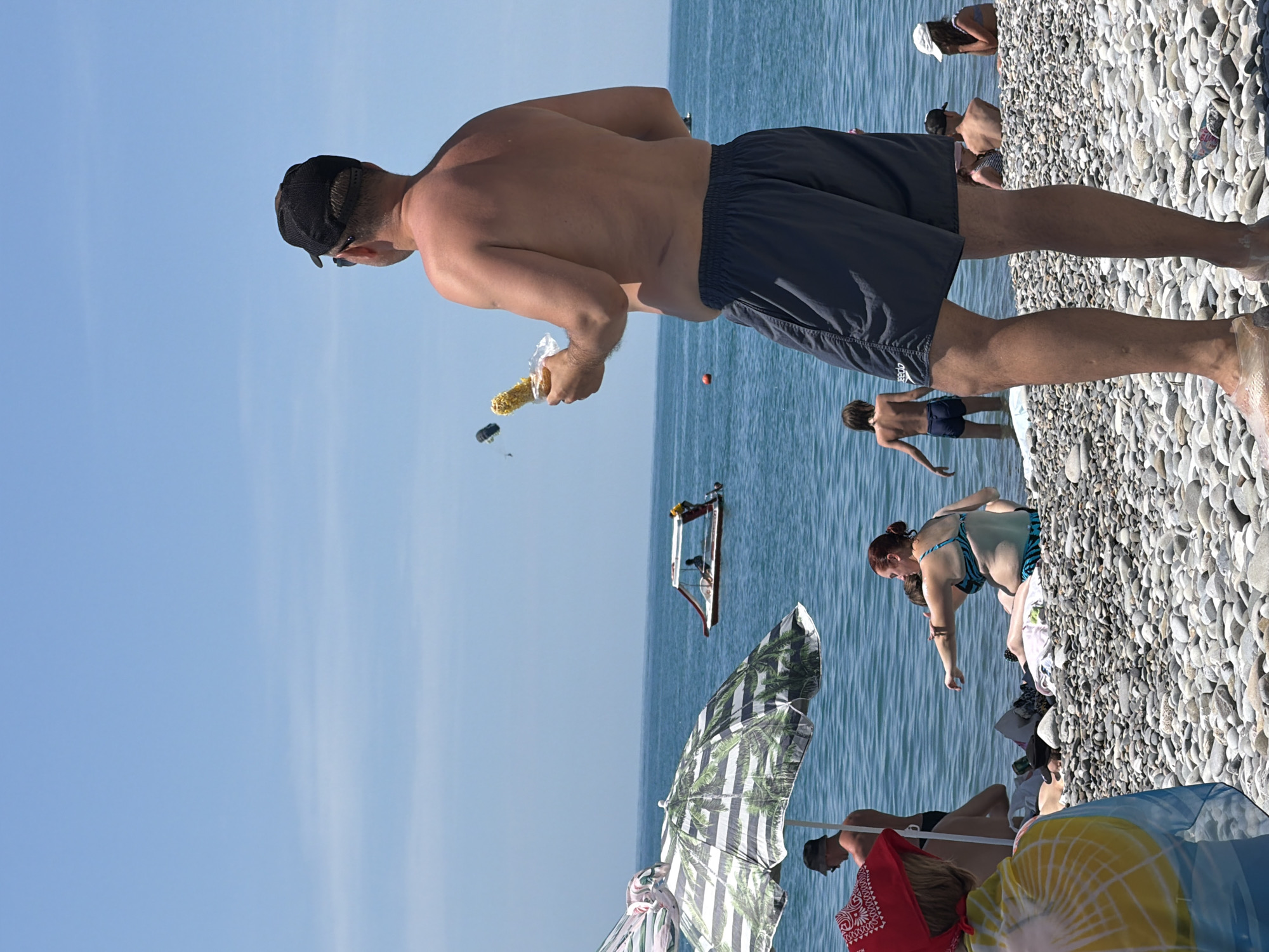 This media file captures a sunny day at Mamedova Shchel', Russia, likely during peak summer. The scene depicts a lively beach with several people enjoying the water and shore.
The primary subject is a man in the foreground, seen from the back, wearing a black cap and dark shorts. He is holding a partially eaten cob of corn in a plastic bag. To his right, in the background, other individuals are visible. A woman in a blue and black bikini is sitting on the pebble beach, and further back, a man is standing in the shallow water. In the distance, a small boat is visible on the calm, blue sea. Beach umbrellas, one with a palm tree pattern, suggest a typical beach setup.
The overall atmosphere is relaxed and vacation-like, indicative of a warm, clear day with bright sunshine. The pebble beach is a distinctive feature of the location. No visible text is present in the media file.