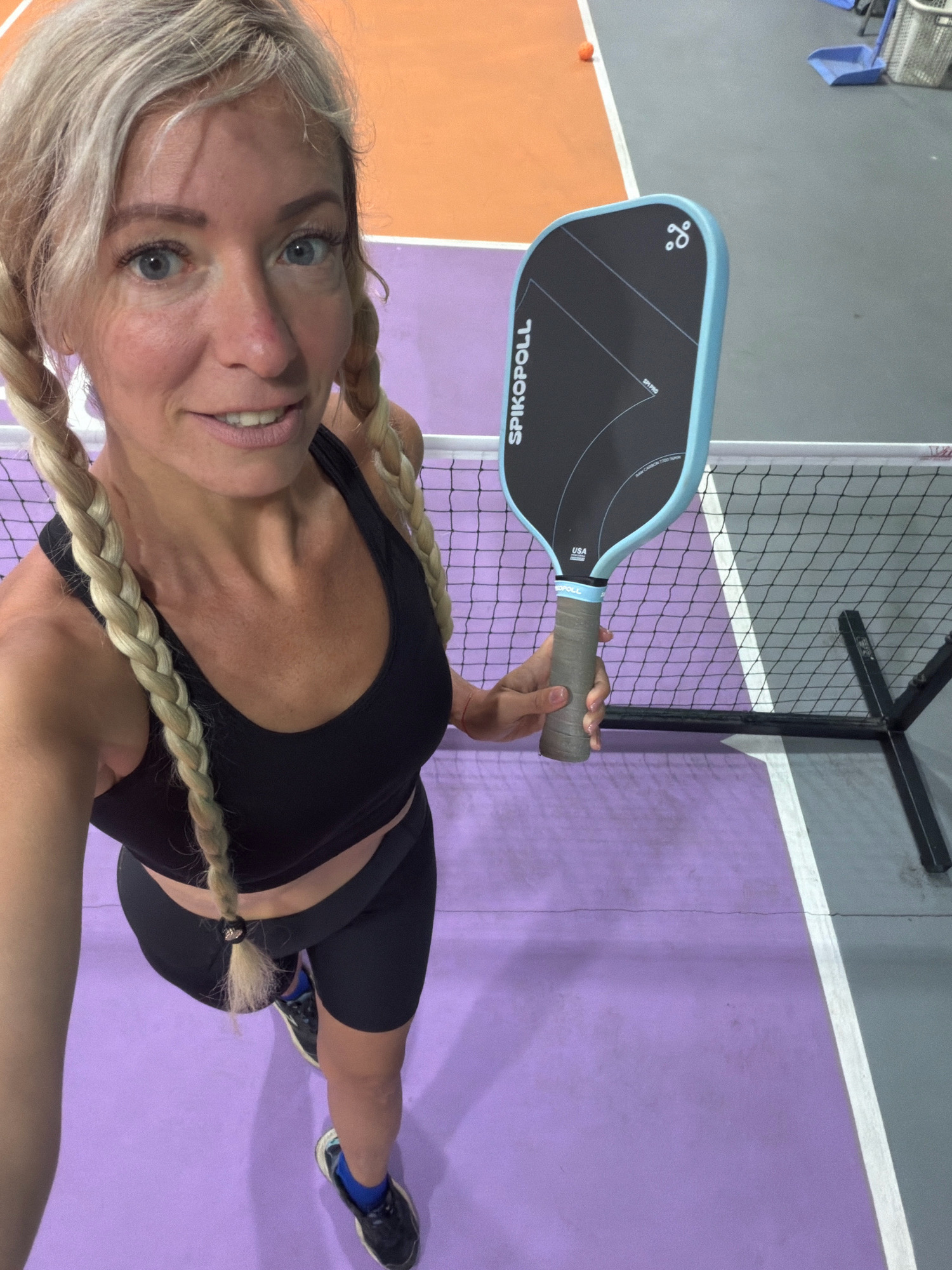 The image shows a woman standing on a pickleball court. She is holding a pickleball paddle and is wearing athletic attire. The court has purple and orange sections, and a net is visible in the background. There is a pickleball on the court. The woman has blonde hair in two braids and is smiling at the camera. The scene is indoors, likely at a sports facility.