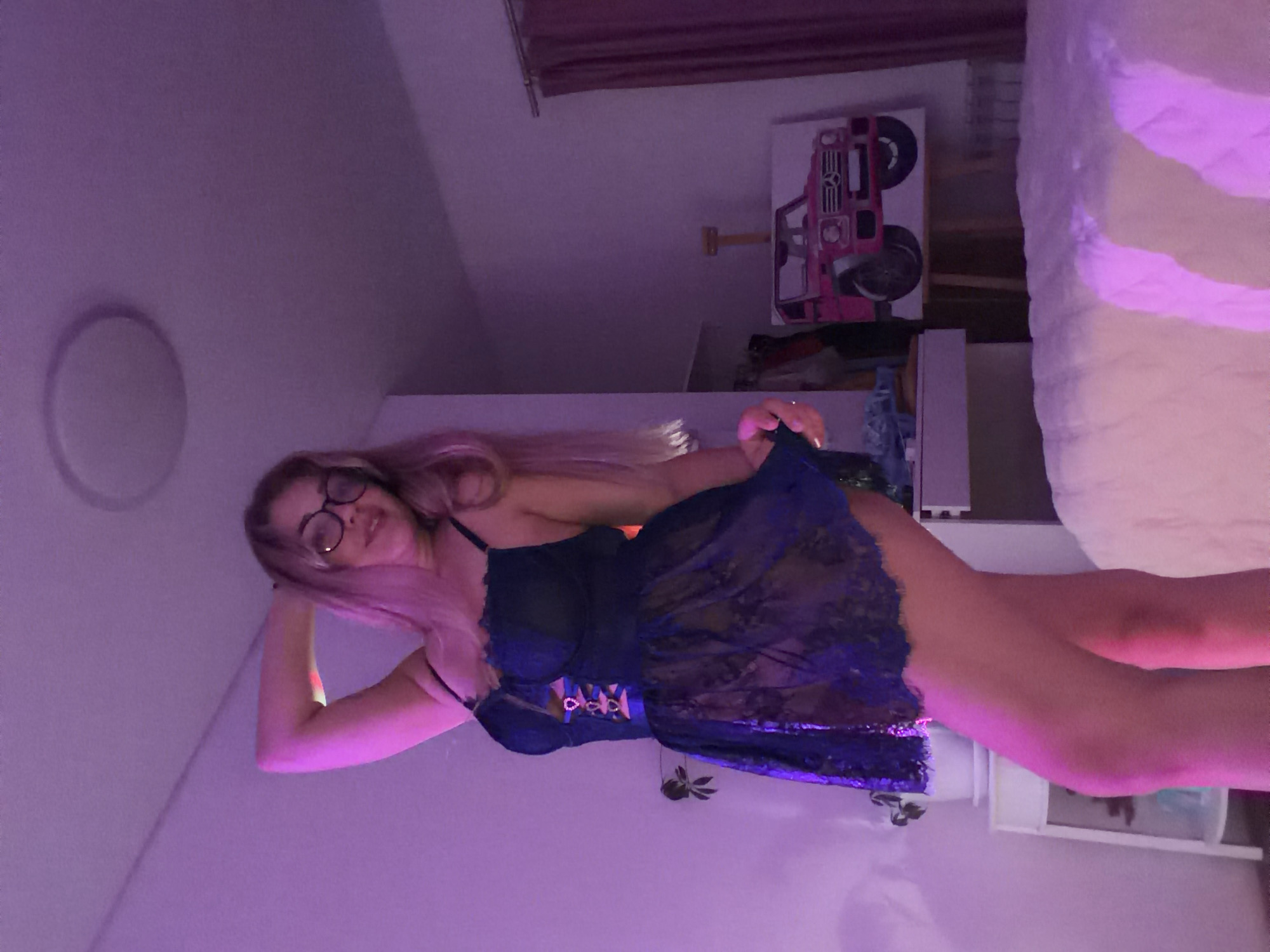 A person with long blond hair and glasses is wearing a blue lingerie-style dress. The person is indoors in a room with purple lighting. The room contains furniture, a painting of a pink vehicle, and a bed partially visible in the upper right corner. There is no indication of a location or city in the image.
