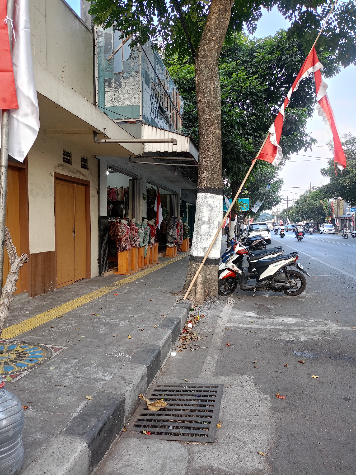 In Batu, Indonesia, a street scene unfolds with a row of shops lining the left side of the image. The shops display various batik clothing items on mannequins and stands. In front of the shops, a sidewalk is marked with a tactile yellow paving strip, indicating accessibility features. A large tree with a trunk painted with white bands stands on the edge of the sidewalk. In front of the tree, a red and white flag is attached to a bamboo pole. A motorcycle is parked near the tree. The street, visible on the right, has traffic, including motorcycles, cars, and a white van. The environment suggests a commercial area with a clear sky above.