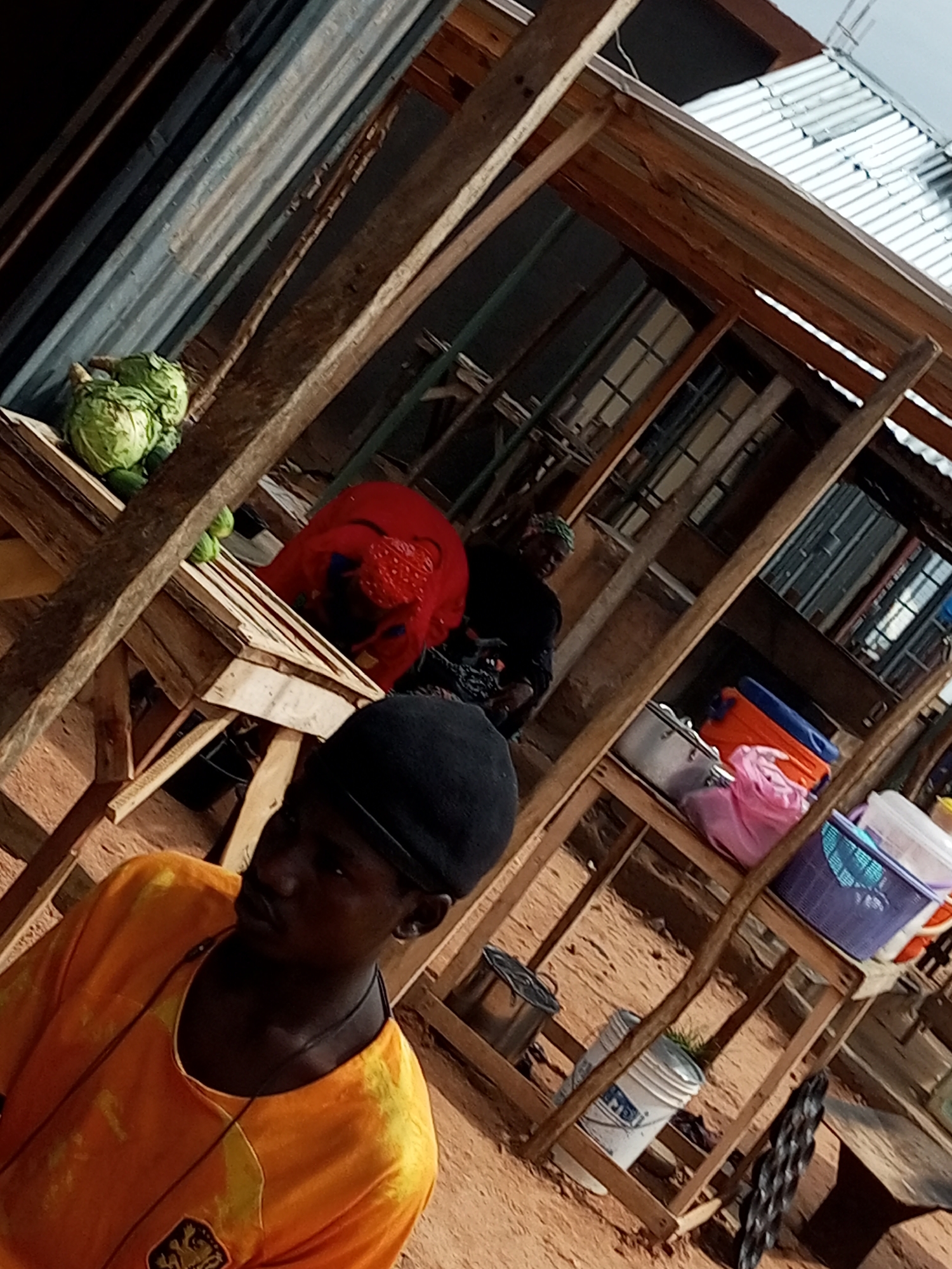 The image captures a scene at an outdoor market in Tempure, Nigeria. In the foreground, a young man wearing an orange t-shirt and a black beanie is looking to his right. Behind him, a wooden stall displays various items. On the top shelf, there are multiple cabbages and what appear to be other vegetables. Further back, two women are present. One woman, wearing a red garment, is partially obscured. Another woman, wearing a dark headscarf, is visible in the background. Shelves of the stall hold several containers, including pots, plastic bags in pink and orange, a blue basket, and white buckets. The market structure is made of wood, with corrugated metal roofing visible in the background. The ground appears to be sandy or dirt. The overall impression is of a busy marketplace environment.