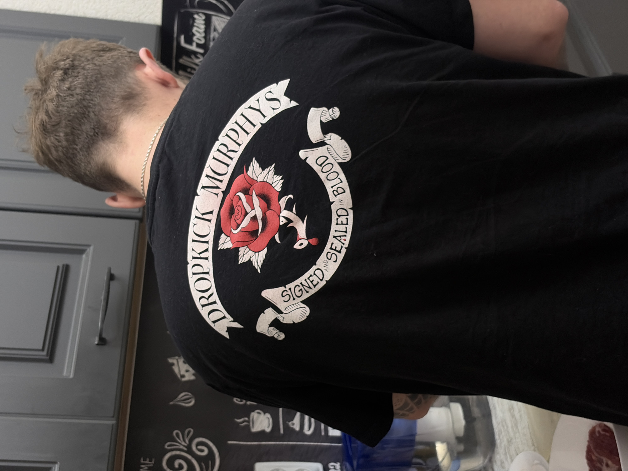 A person is depicted from behind, wearing a black t-shirt with a prominent graphic design, likely standing in a kitchen. The individual has short, light brown hair and is wearing a thin silver chain around their neck. The t-shirt's design features a large red rose with green leaves and thorns, encircled by a white banner.
The banner displays text in a stylized font. The upper arc reads "DROPKICK MURPHYS", and the lower arc, which is inverted, reads "SIGNED AND SEALED IN BLOOD". Below the rose, a small, stylized anchor or similar design is visible.
The background shows grey kitchen cabinets with dark handles on the left. To the right, behind the person, is a dark surface, possibly a decorative kitchen mat or wall hanging, with white chalk-like drawings. Partially visible text on this surface, inverted in orientation, includes "Milk Foame" and other small icons and numbers such as "ME", "40", and "22", suggesting a coffee or kitchen theme. The scene appears to be indoors with ambient lighting. While the provided context indicates Saint Petersburg, Russia, the image itself does not contain specific visual cues to confirm this location.