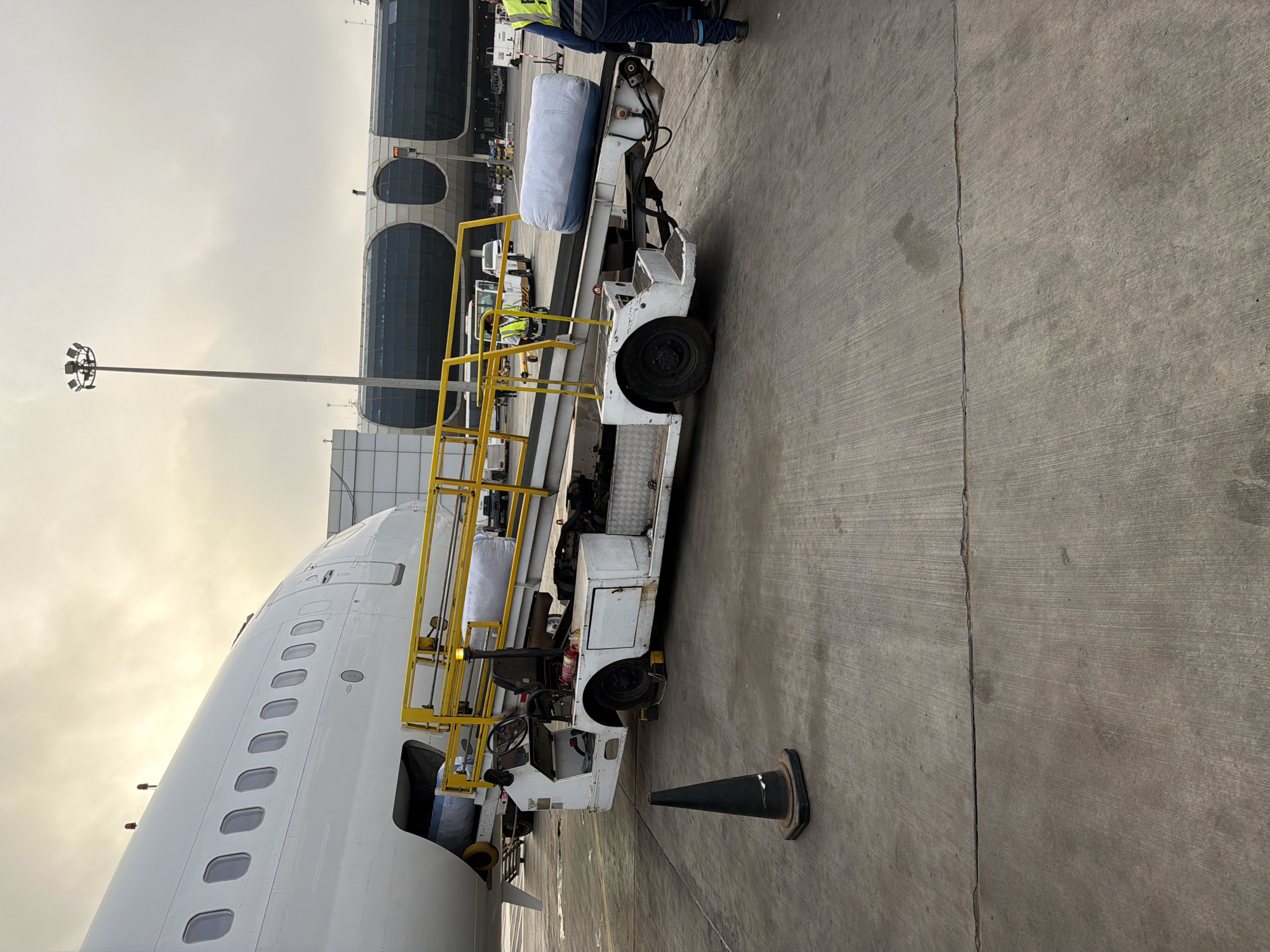 The image shows the exterior of an airport in Communauté rurale de Diass, Senegal. A white passenger airplane is parked on the tarmac. A white luggage carrier with yellow railings is parked alongside the airplane. A black traffic cone stands nearby on the concrete. An airport terminal is visible in the background. The sky is overcast. An airport light pole is visible in the background. A person wearing a neon yellow vest is partially visible on the luggage carrier.