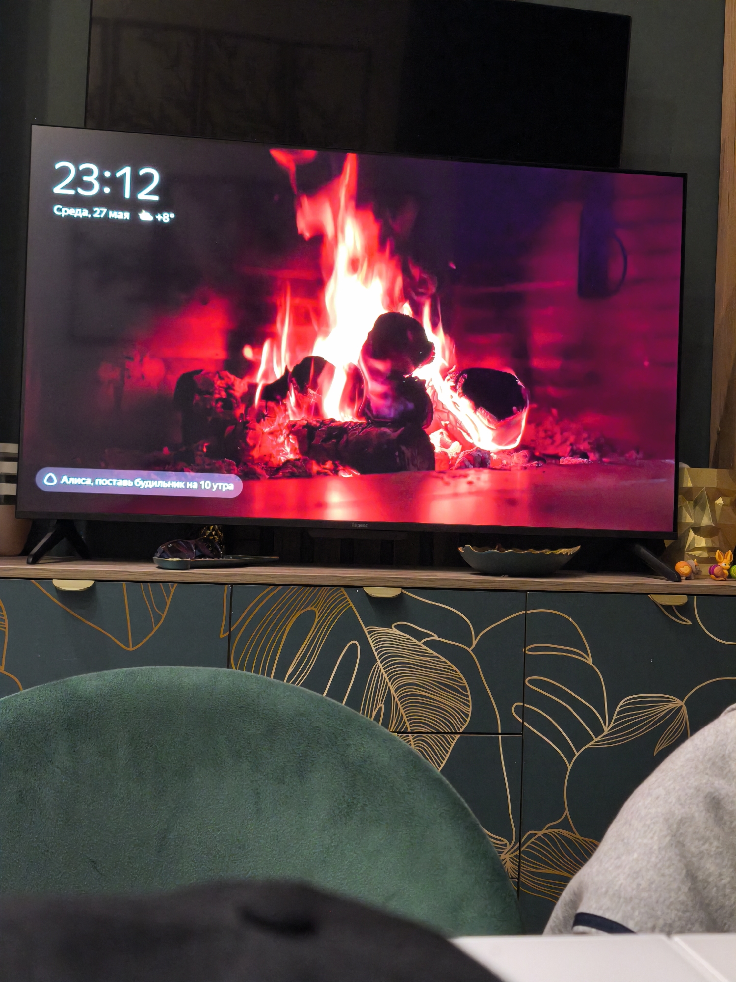 Here is a forensic description of the image:
The image depicts an indoor scene dominated by a large flat-screen television displaying a looping video of a roaring fireplace with flames. The time displayed in the upper left corner of the screen indicates "23:12", and the date is specified as "Среда, 27 мая", along with a temperature of "+8°". Below the time, a banner appears to read "Алиса, поставь будильник на 10 утра", which translates to "Alice, set an alarm for 10 a.m.", alongside the "Яндекс" logo.
The TV rests on a wooden cabinet with dark green doors decorated with gold botanical line art. A pair of sandals, a small bowl, a gold-toned geometric object resembling a face, and a couple of small animal figurines are visible on top of the cabinet.
In the foreground, part of a green velvet chair is visible, along with a partially visible person wearing a grey sweater. The setting appears to be within a home environment.