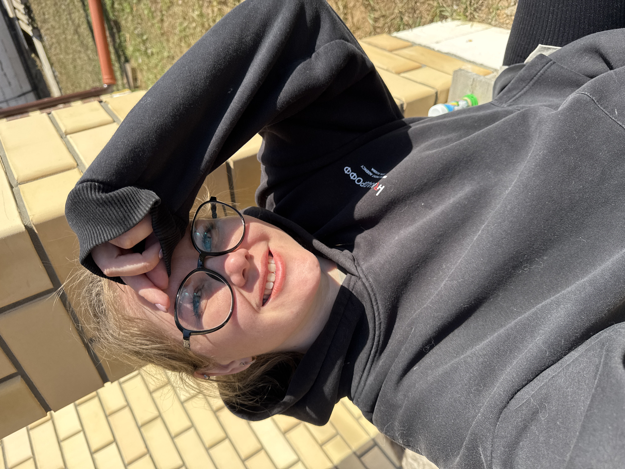 A person is lying on a stone or brick surface outdoors, wearing a black hooded sweatshirt and glasses. The person is smiling. The setting appears to be an outdoor area in Gornyi, Dvurechenskoe S/P, Russia, as indicated by the image tags. There are brick or stone structures visible in the background. A small bottle with a green cap is also visible.