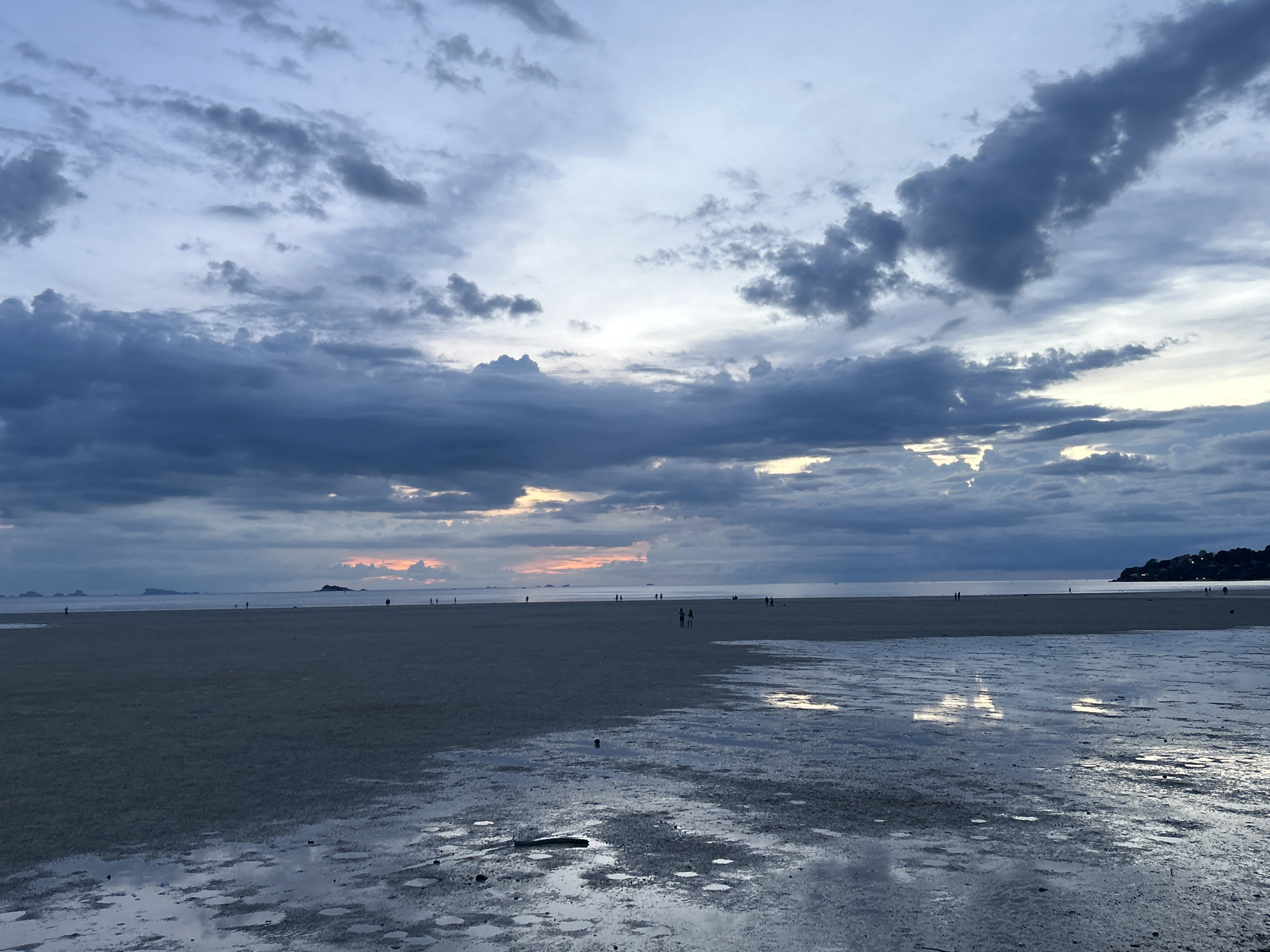 The image depicts a low tide beach scene at dusk on Koh Phangan, Thailand. The sky is filled with dramatic, dark clouds, with breaks allowing soft, diffused light to illuminate the horizon where a faint pink and orange glow suggests the setting sun. The foreground is a wet, sandy expanse with numerous shallow pools reflecting the sky and clouds. In the middle ground, a few silhouetted figures can be seen walking on the beach, some in small groups. In the distance, islands or landmasses are visible on the horizon. The overall atmosphere is serene and somber, characteristic of twilight on a tropical island.