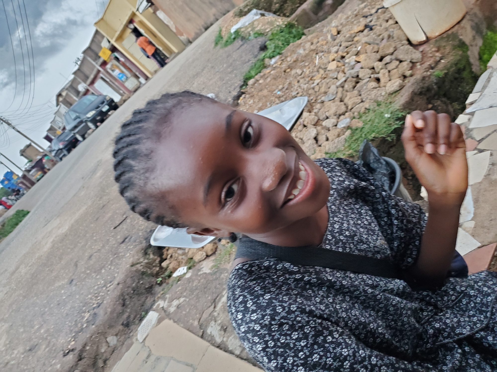 A young girl with braided hair smiles at the camera, her head tilted to the side. She wears a dark, floral-patterned dress. Her right hand is visible in the foreground, raised as if gesturing or about to touch the camera. The background depicts a street scene in Jos, Nigeria. Buildings line the street, some with corrugated iron roofs. A black car is parked on the left side of the road, and the ground appears to be a mix of asphalt and rough terrain with scattered rocks and some patches of green grass. The sky is overcast, suggesting cloudy or possibly rainy weather. The overall impression is a candid, informal portrait captured outdoors in a bustling, everyday setting.
