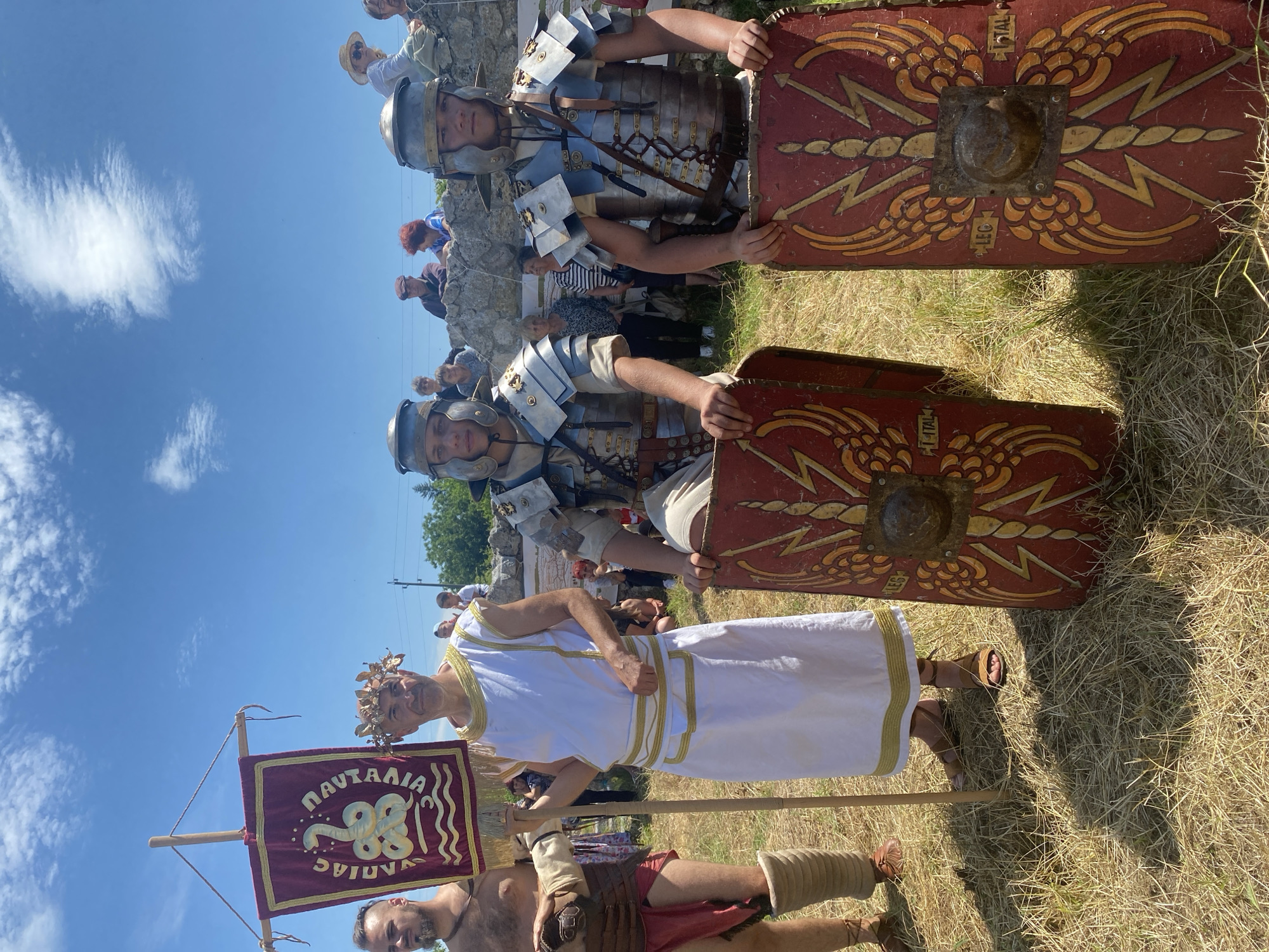 This photograph depicts a historical reenactment event in Gigen, Bulgaria, under a clear blue sky with scattered white clouds. Four individuals in period attire stand prominently in the foreground on dry, yellowish grass. On the far left, a bare-chested male with a beard wears leather armor straps, a red loincloth, and sandals. Adjacent to him, an older male with a golden leaf wreath on his head wears a white tunic with gold trim and holds a maroon standard vertically. The standard features a gold coiled snake above wavy lines, with Greek text reading "SYNAIAΣ" vertically on the left and "NAVTALIA" on the right. To the right of the standard bearer, two younger males, dressed as Roman legionaries, wear helmets and lorica segmentata armor over tunics. Each holds an upright, rectangular scutum (shield) on the ground. The shields are red with gold winged lightning bolt designs, a central metallic boss, and the inscriptions "LEG" and "I ITAL". In the background, multiple spectators are visible behind a low stone wall or ruins, observing the scene. Trees and a power line are discernible in the distant background.