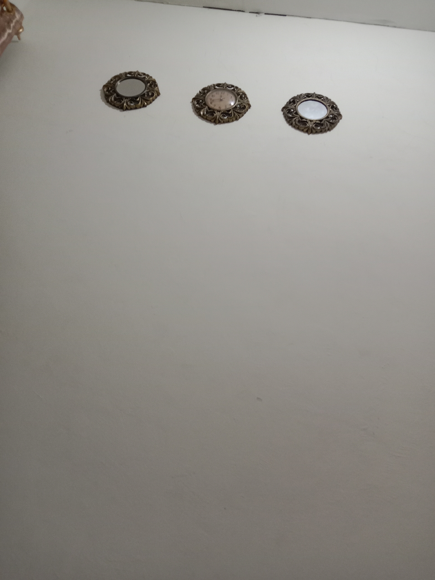 A wall with three decorative circular objects is visible. The object on the left is a small mirror with an ornate, gold-colored frame. To its right is a clock with a similar gold-colored frame, featuring Roman numerals and a light-colored face. Further to the right is another small mirror with an ornate, gold-colored frame. The wall is painted a light, neutral color. The lighting suggests an indoor environment, possibly a room in Brangsong, Indonesia. There are no people or interactions depicted in the image.