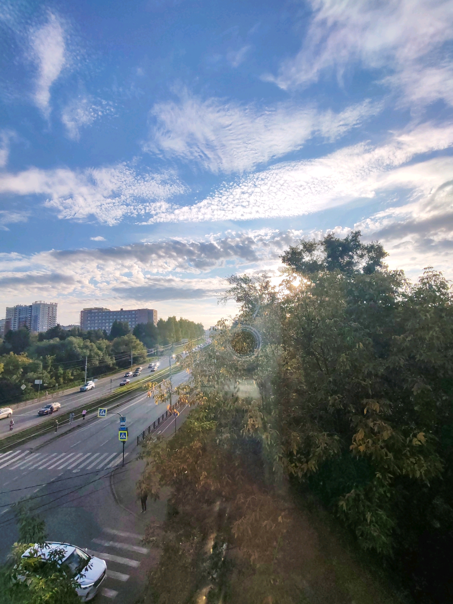 The image depicts a city street scene in Izhevsk, Russia, under a bright, partly cloudy sky. In the foreground, a white car is parked on the left side of the frame, partially obscured by foliage. Adjacent to it is a crosswalk. Further down the street, a multi-lane road carries traffic, with several cars visible. Pedestrians can be seen walking on the sidewalk and crossing the street. Traffic lights and pedestrian crossing signs are present. In the background, apartment buildings and lush green trees line the street, with the sun setting or rising behind them, casting a warm glow and creating lens flare. The overall impression is of a typical urban environment during daylight hours.