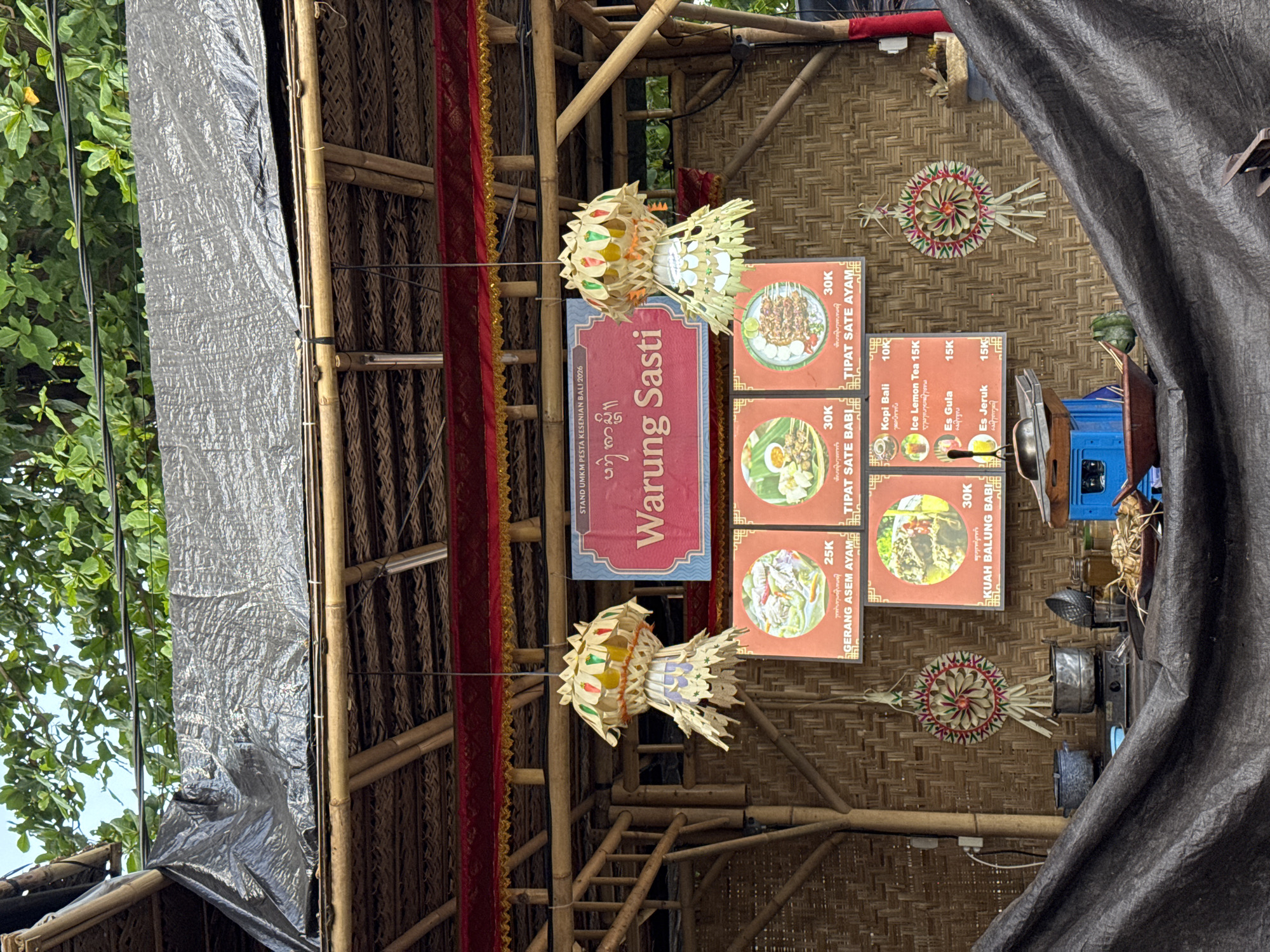 The image depicts an outdoor food stall named "Warung Sasti" in Denpasar, Indonesia. The stall is constructed with bamboo and palm fronds, with a tarp covering the top. A red and gold banner hangs above the stall, and decorative paper lanterns are suspended from the ceiling. Several framed menu items are displayed on the bamboo wall, showcasing food and drinks with prices in Indonesian Rupiah. In the foreground, a table is set up with various cooking utensils, pots, and prepared food. The overall scene suggests a bustling local eatery, likely serving traditional Indonesian cuisine.