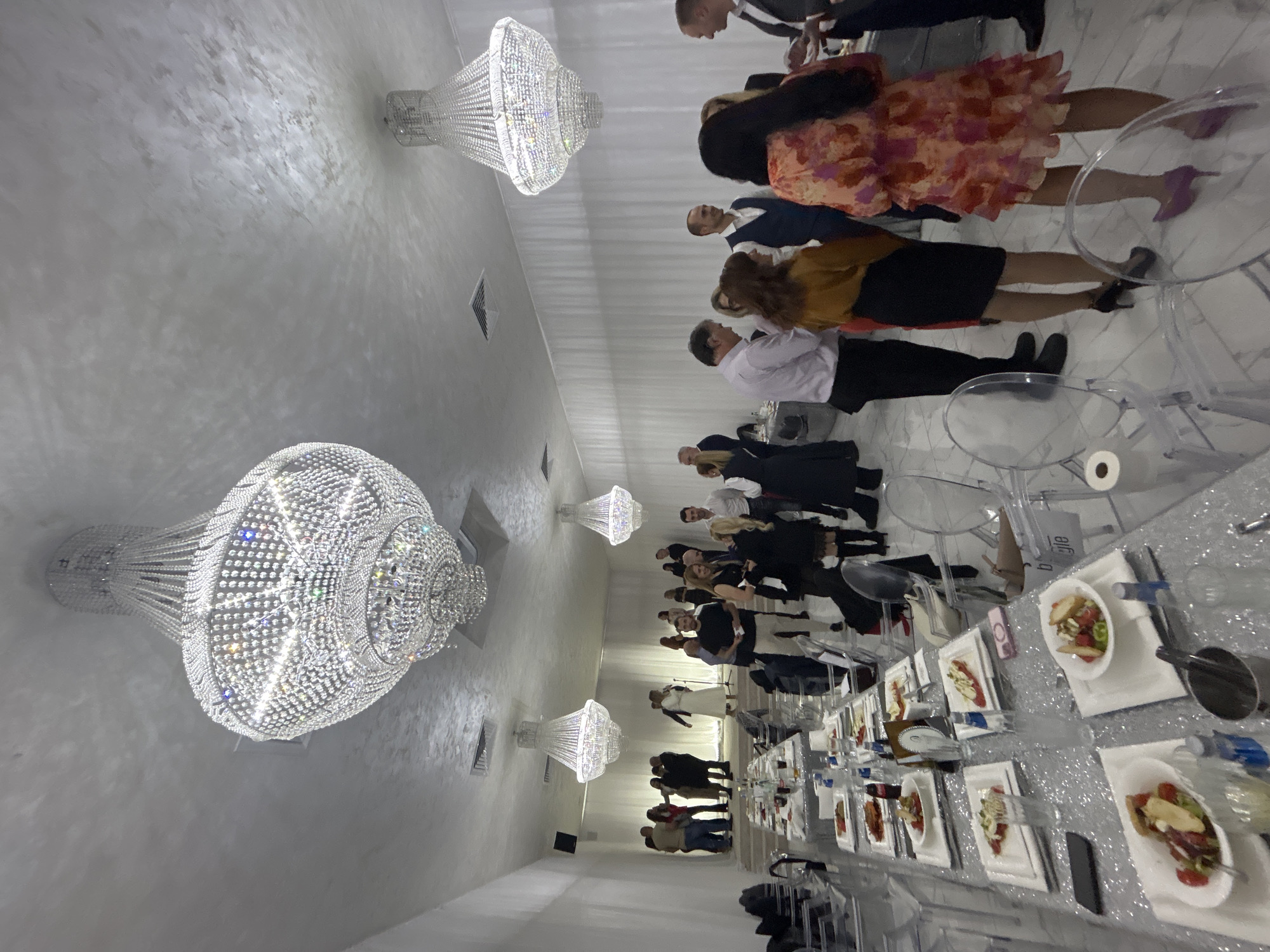The image captures an event at a banquet hall in Plovdiv, Bulgaria. The venue is brightly lit, with multiple crystal chandeliers illuminating the space. Guests are dressed in formal attire, some standing near the tables and others seated. Tables are arranged in rows and covered with glittery tablecloths. The tables are set with plates of food, cutlery, glasses, and water bottles. People are interacting, talking, and enjoying the event. The hall is white and appears modern.