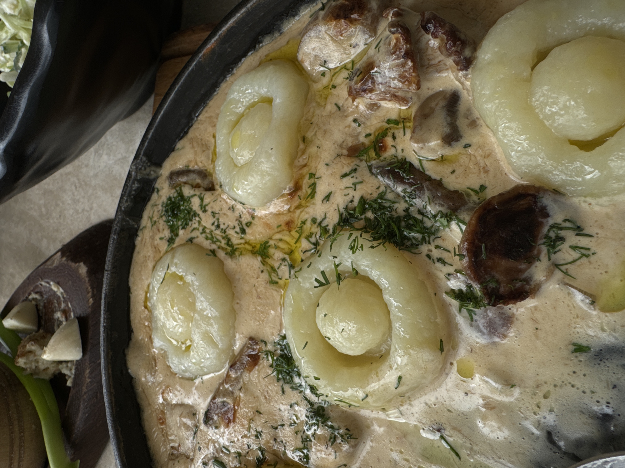 A close-up, overhead shot shows a portion of a meal in a dark, round pan filled with a creamy sauce. The dish contains several round, pale dumplings, each with a small ball of dough in the center, surrounded by a ring of dough. Sliced mushrooms are visible throughout the sauce, along with chopped green herbs, likely dill. The pan is set on a textured surface, possibly a table, and part of a dark bowl containing a green salad is visible in the upper right corner. In the upper left, a wooden board holds a few pieces of bread and two cloves of garlic, with a green onion stalk resting on top. The image does not contain any recognizable landmarks or elements that would specifically place it in Polianytsia, Ukraine; it depicts a prepared food item.