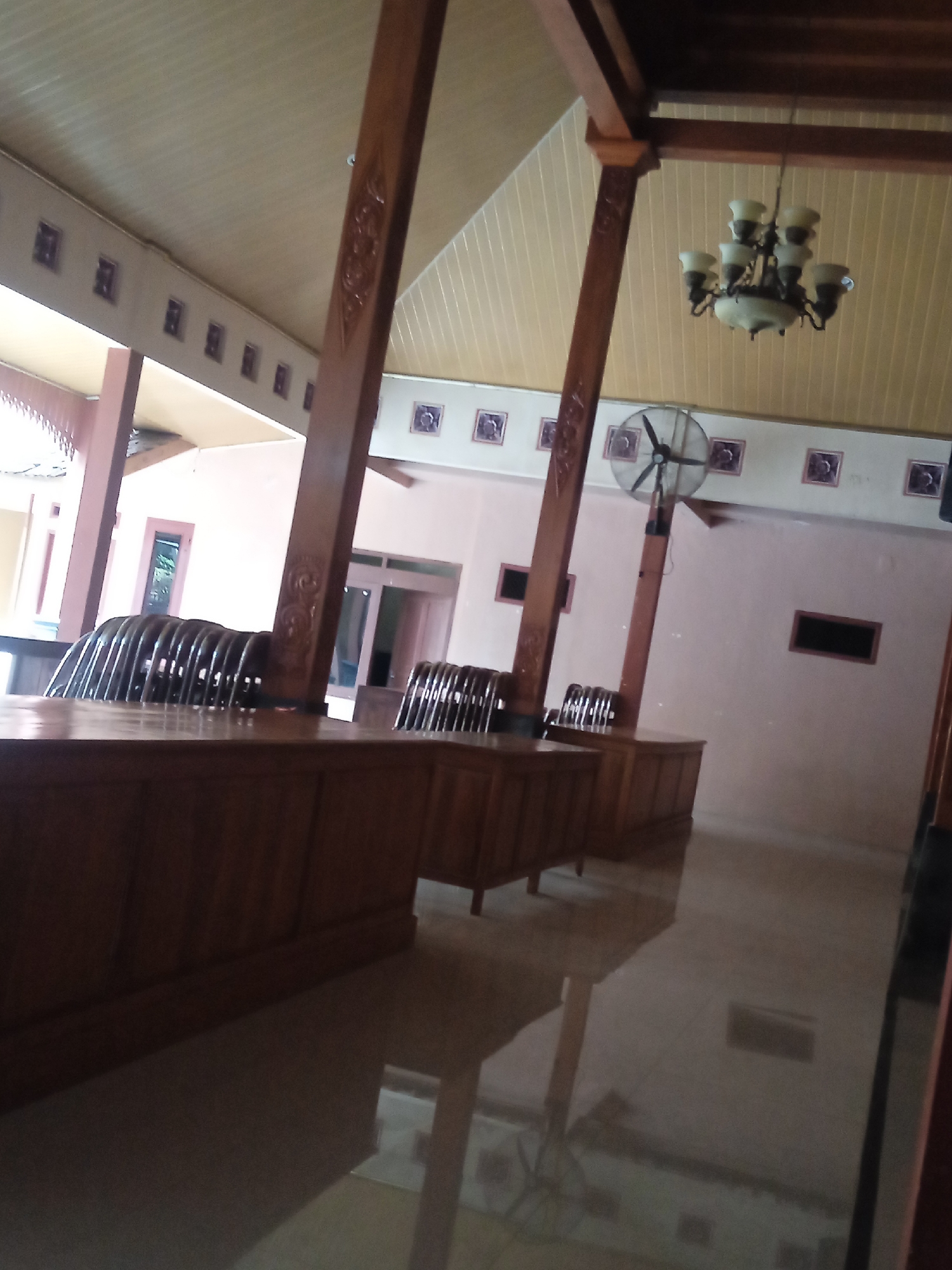 The image shows the interior of a building with a wooden roof structure and light-colored walls. In the foreground, there are long, wooden counters or tables. Behind these, rows of chairs with a distinctive curved, woven back are visible. Further back, a ceiling fan is mounted on a wooden pole, and a chandelier hangs from the ceiling. The walls are decorated with framed squares. The floor is polished and reflects the overhead lighting and the wooden structures. The scene suggests an indoor waiting area or reception space, possibly in a community building or office in Sugihan, Indonesia.