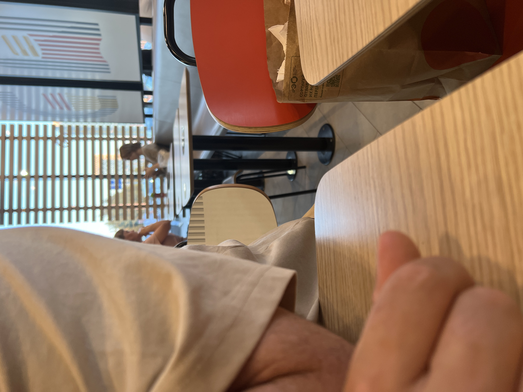 This media file depicts a close-up, rotated view of a person's hand and arm resting on a light-colored wooden table. The setting appears to be indoors, likely a restaurant or cafe, with other tables and chairs visible in the background. A red chair with a black frame is prominent, along with a paper bag from a fast-food establishment, featuring Cyrillic text and a QR code. The background also shows vertical blinds, suggesting a window. In the distance, through the blinds, a person can be seen sitting at a table. The overall lighting suggests daytime. The image is cropped and rotated, emphasizing the texture of the table and the fabric of the person's clothing. There is no indication of the specific time of day, weather conditions, or overt emotions. The visible text on the paper bag includes Cyrillic characters, consistent with a Russian location.