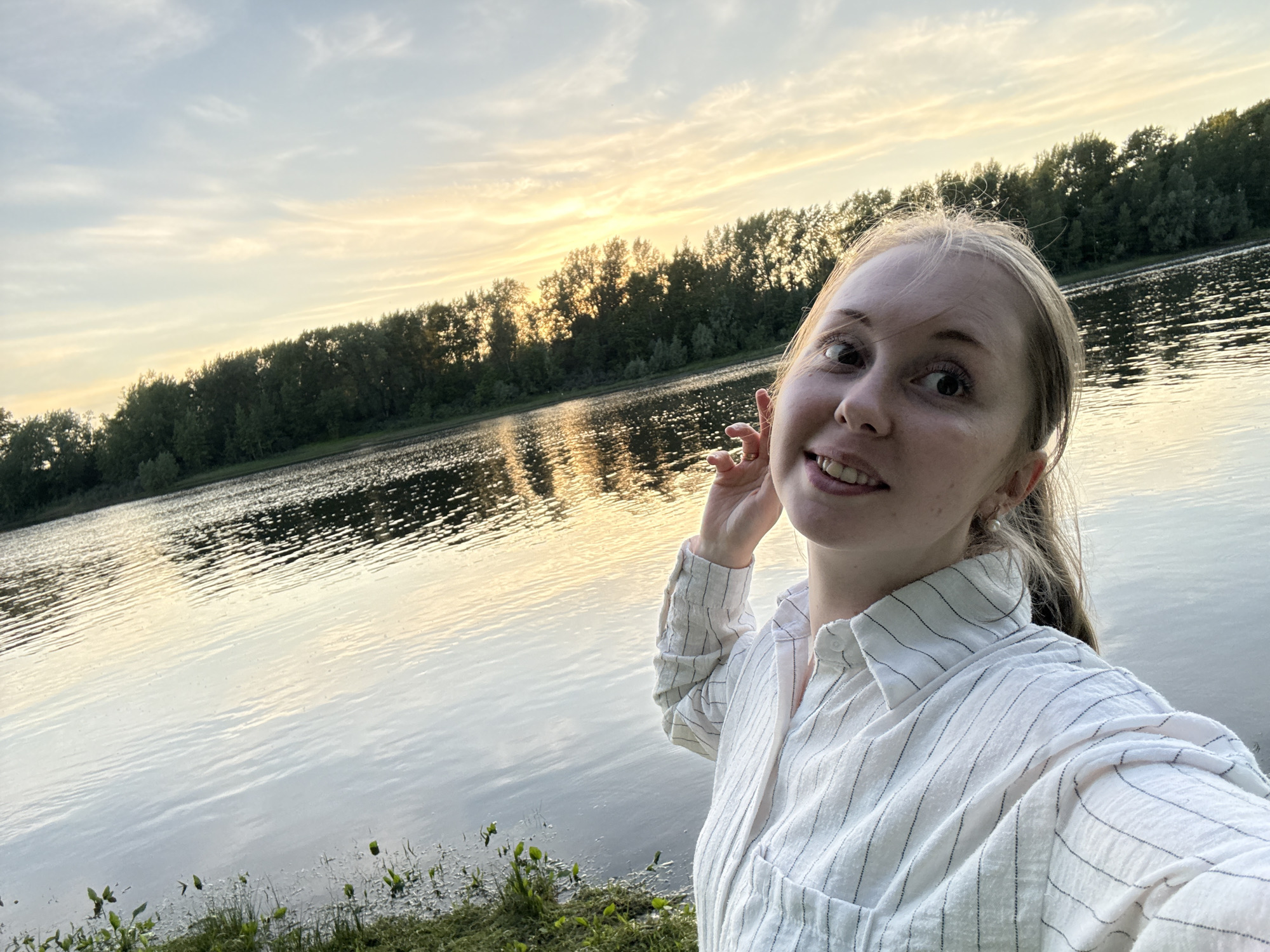 A young woman with blonde hair takes a selfie by a wide river during sunset. The sky is a soft mix of blue and pale yellow, with wispy clouds. A dense line of dark green trees runs along the opposite bank of the river, silhouetted against the colorful sky. The water is calm, reflecting the warm hues of the sunset in shimmering streaks. The woman is smiling and looking at the camera, with one hand touching her ear. She is wearing a white, pinstriped button-down shirt. The foreground shows a grassy bank with some small green plants.